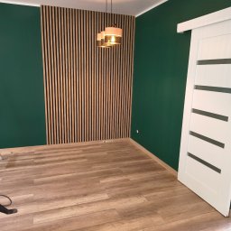 Artur Rzemiszke Usługi Remontowo-Wykończeniowe - Nowoczesne wnętrze z zielonymi ścianami, drewnianą lamelą na ścianie i przesuwnymi białymi drzwiami. Widoczna drewniana podłoga i wisząca lampa.