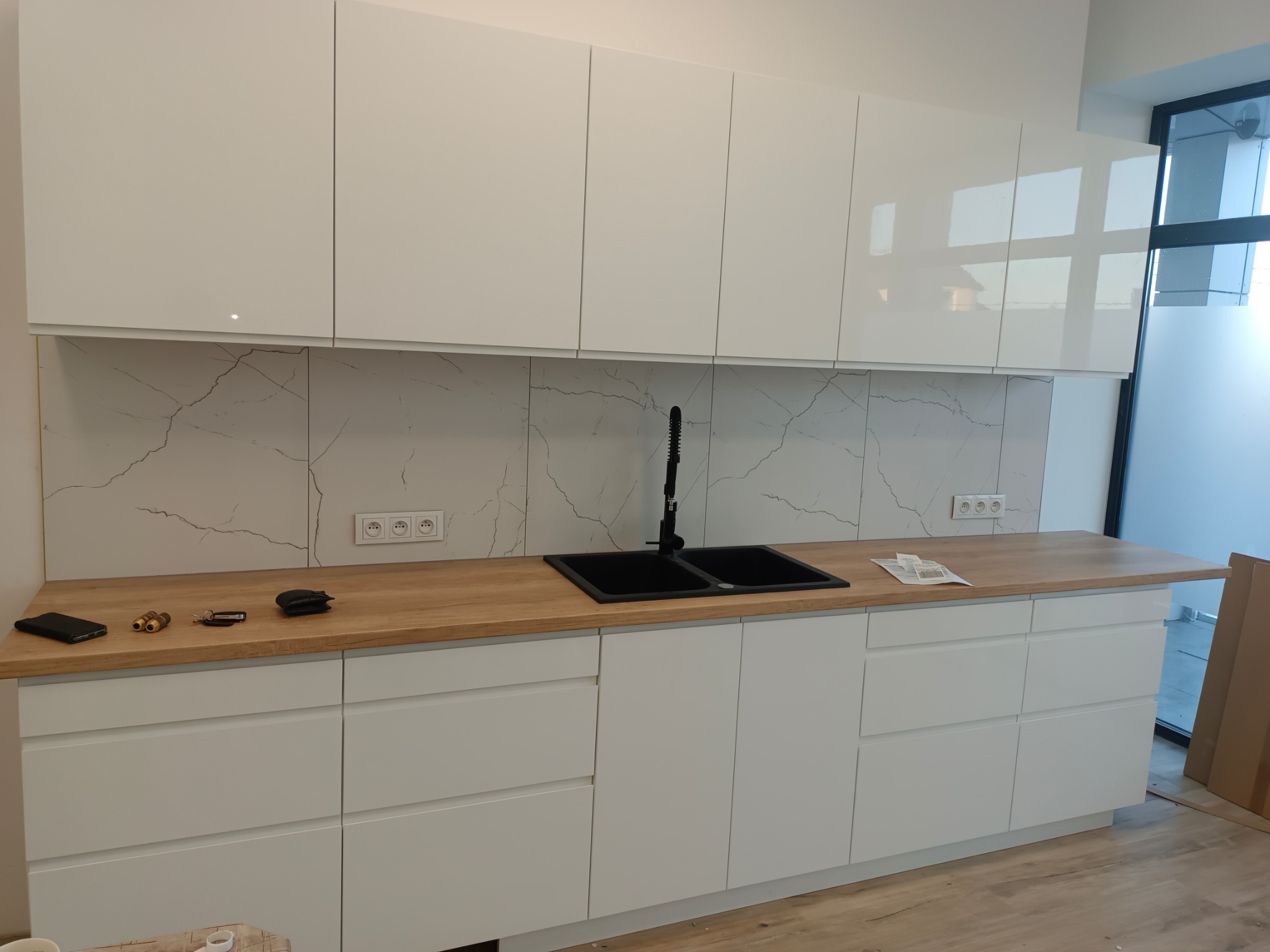 Biała, nowoczesna kuchnia z drewnianym blatem i czarnym zlewem. Fronty szafek na wysoki połysk, marmurowe płytki na ścianie. Widoczne akcesoria na blacie.