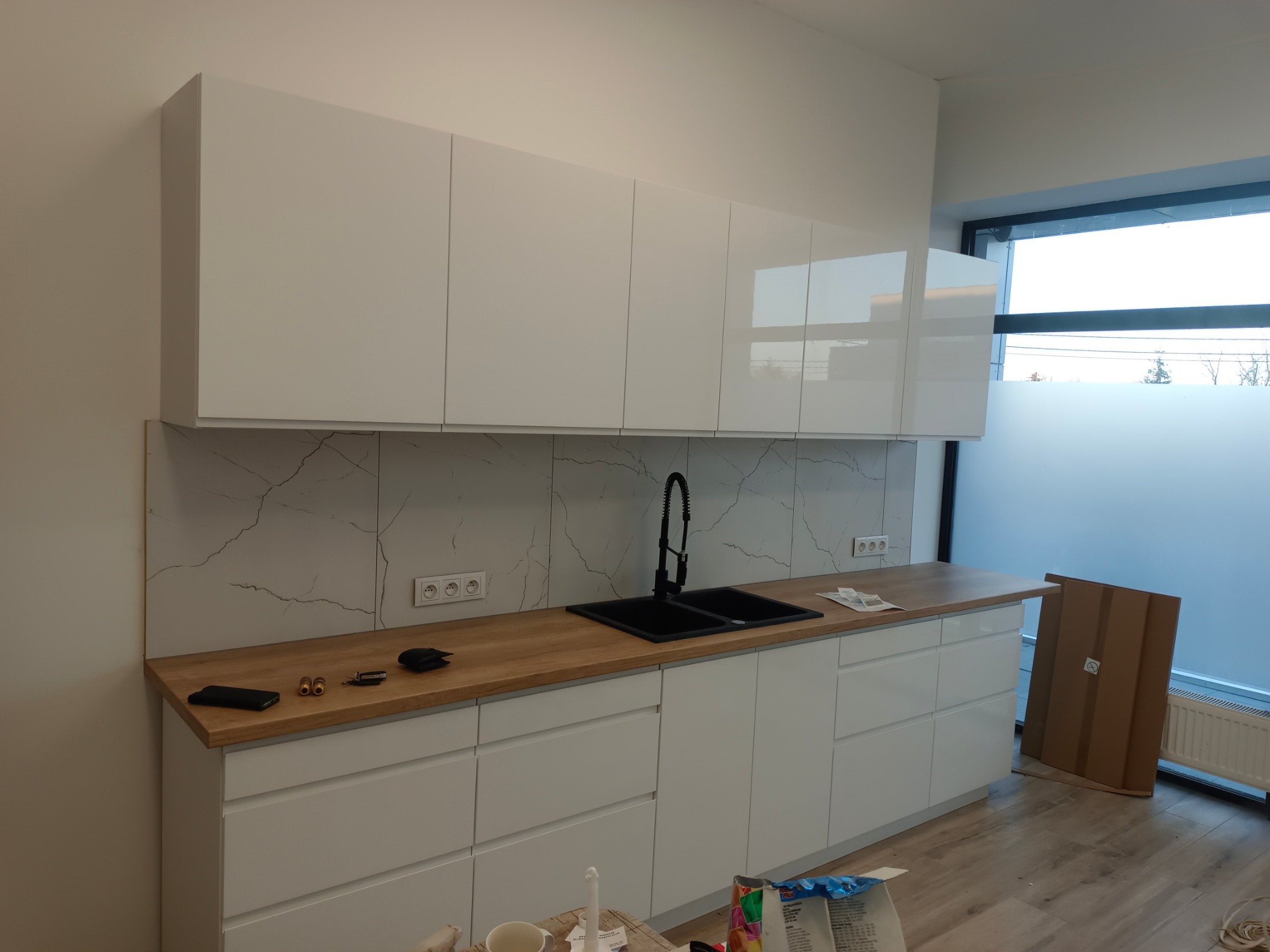 Nowoczesna, biała kuchnia z drewnianym blatem i czarnym zlewem. Marmurowe płytki na ścianie. Minimalistyczny design, widok na okno.