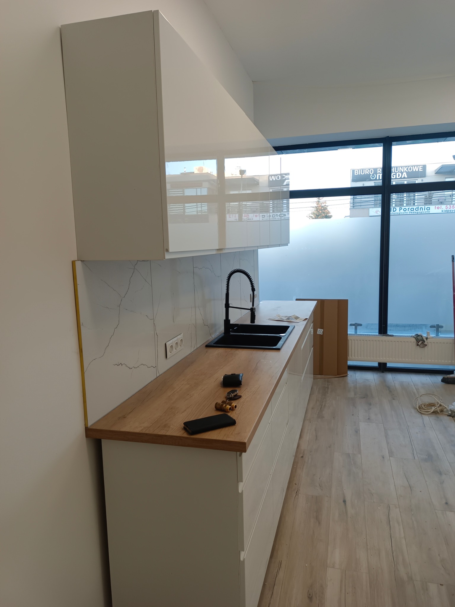 Nowoczesna, jasna kuchnia z drewnianym blatem i marmurową ścianą. Widoczny zlewozmywak z czarną armaturą i elementy instalacji hydraulicznej na blacie.