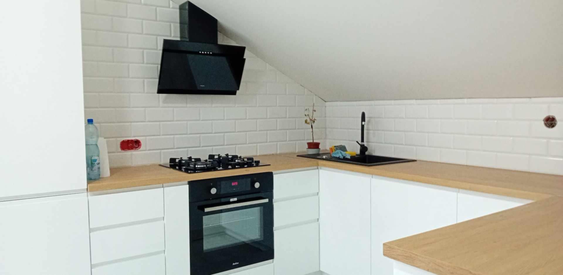 Nowoczesna kuchnia na poddaszu: białe szafki, drewniany blat, czarny okap i płyta gazowa. Ściany wyłożone białymi płytkami imitującymi cegłę.