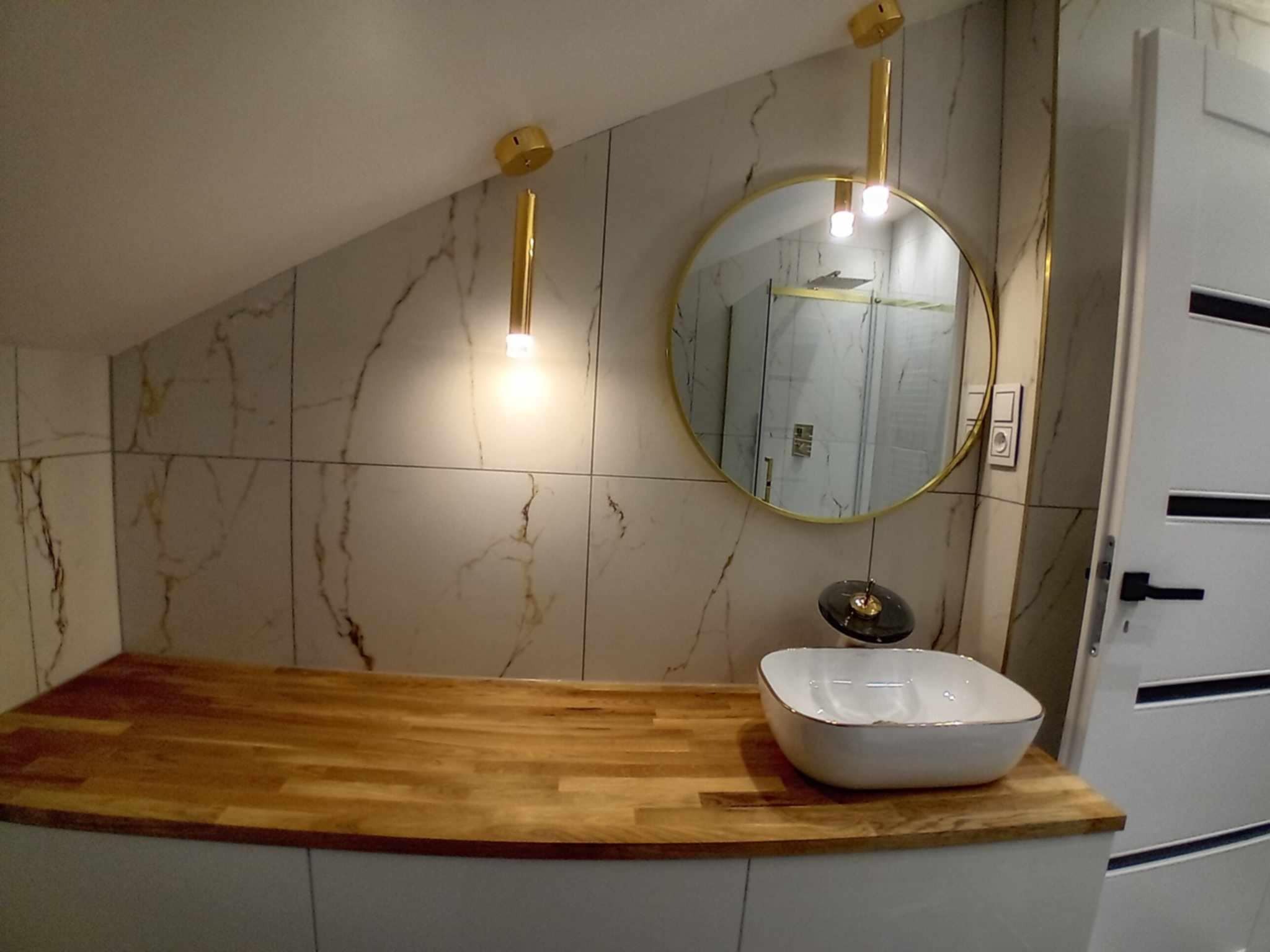 Nowoczesna łazienka na poddaszu: marmurowe płytki, drewniany blat z białą umywalką, okrągłe lustro w złotej ramie i wiszące lampy. Widoczna biała drzwi z czarną klamką.