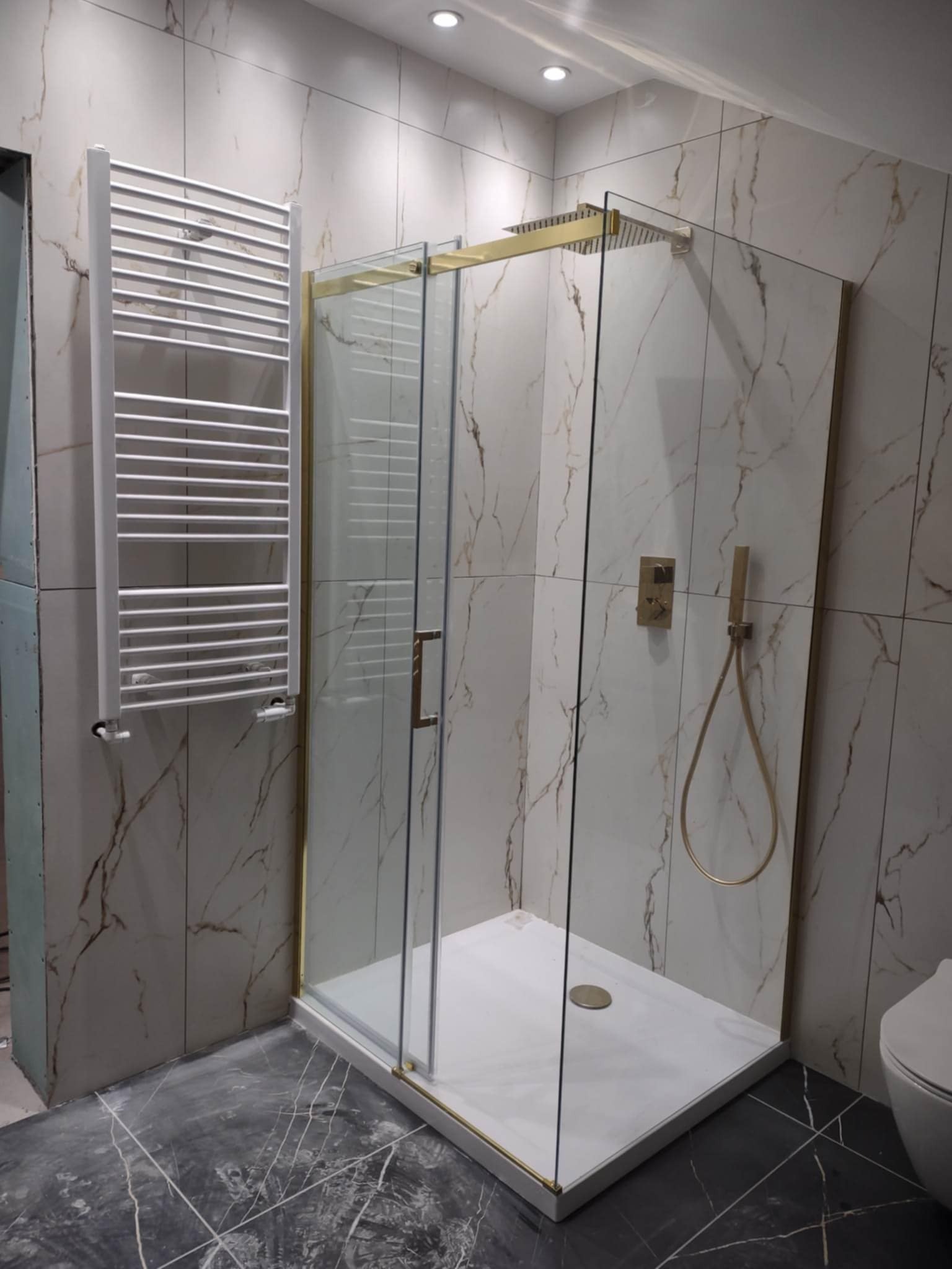 Wykończona łazienka: kabina prysznicowa ze złotymi detalami, grzejnik drabinkowy i marmurowe płytki ścienne. Eleganckie wnętrze po remoncie.