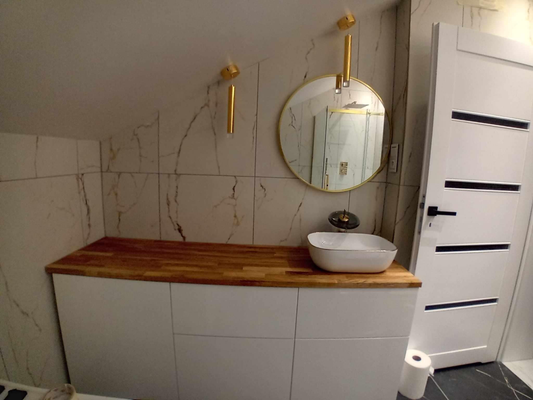 Nowoczesna łazienka w skosach: biała szafka z drewnianym blatem, okrągłe lustro i designerskie lampy na tle marmurowych płytek. Widoczna biała, minimalistyczna umywalka.