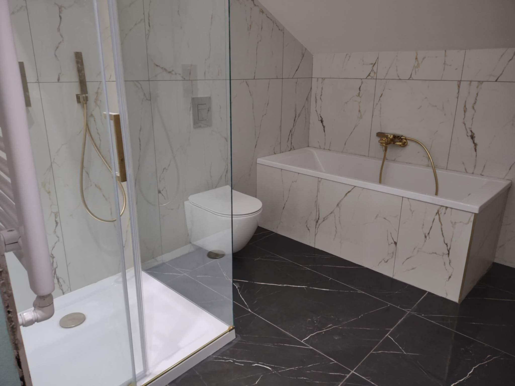Wykończona łazienka w bieli i czerni: kabina prysznicowa ze złotymi akcentami, toaleta podwieszana, wanna obudowana płytkami imitującymi marmur, czarna marmurowa podłoga.