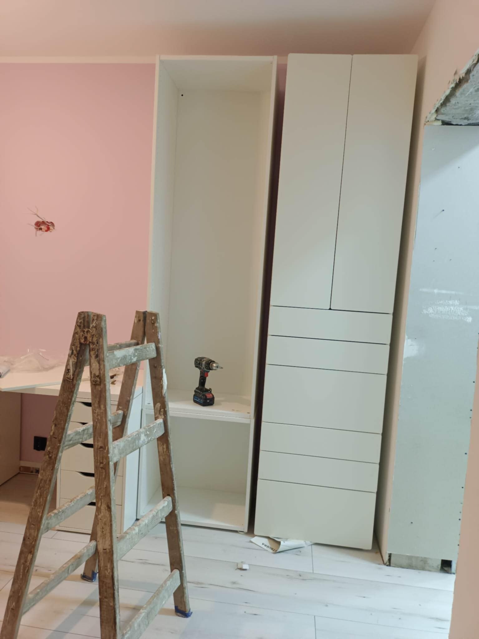 Montaż białych szaf w pokoju z różową ścianą. Widoczna drabina, wiertarka na półce. Podłoga z paneli. Częściowo widoczna płyta karton-gips.