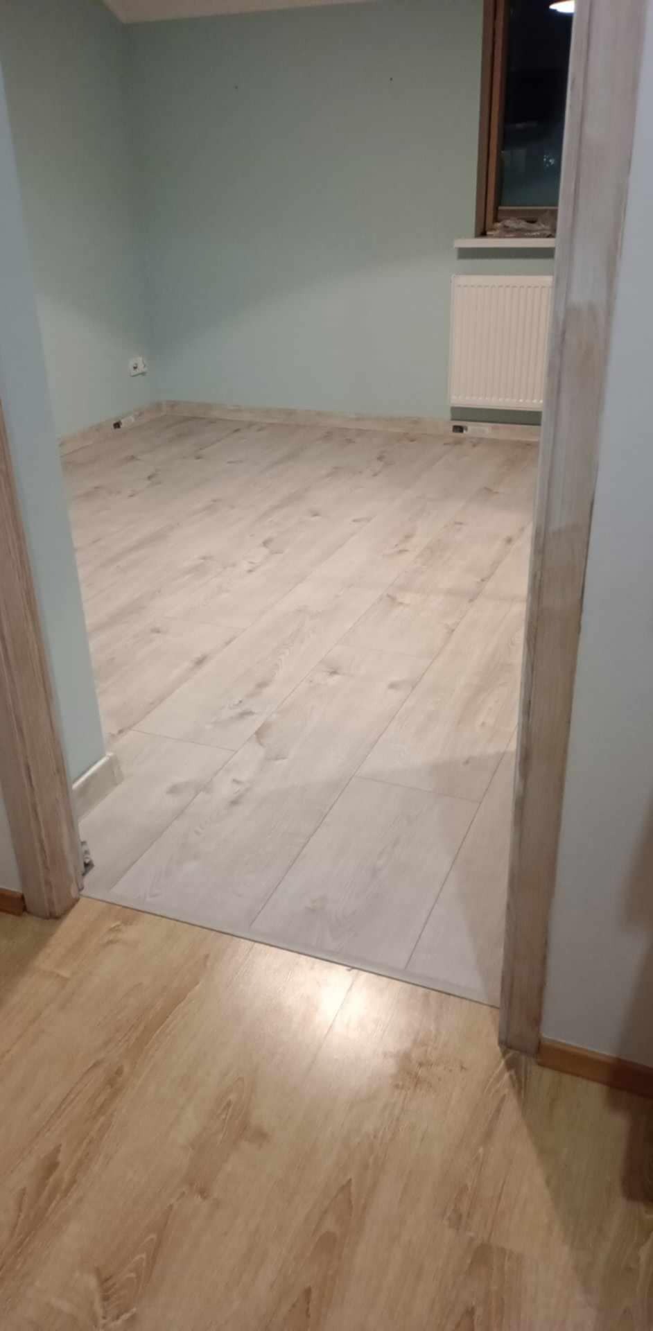 Jasne panele podłogowe ułożone w pokoju z pomalowanymi na jasnozielono ścianami, widok z progu drzwi. Grzejnik pod oknem.