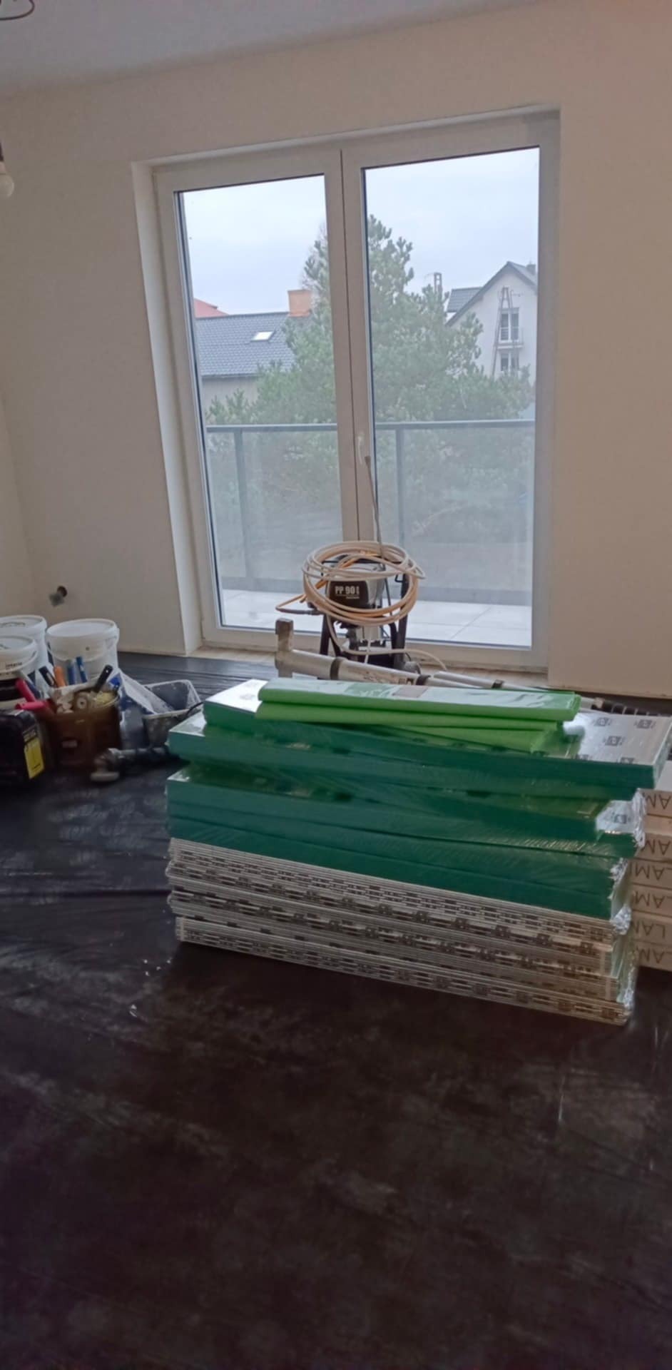 Przygotowanie do montażu paneli podłogowych: narzędzia, podkład i panele na czarnej folii w jasnym wnętrzu z oknem balkonowym.