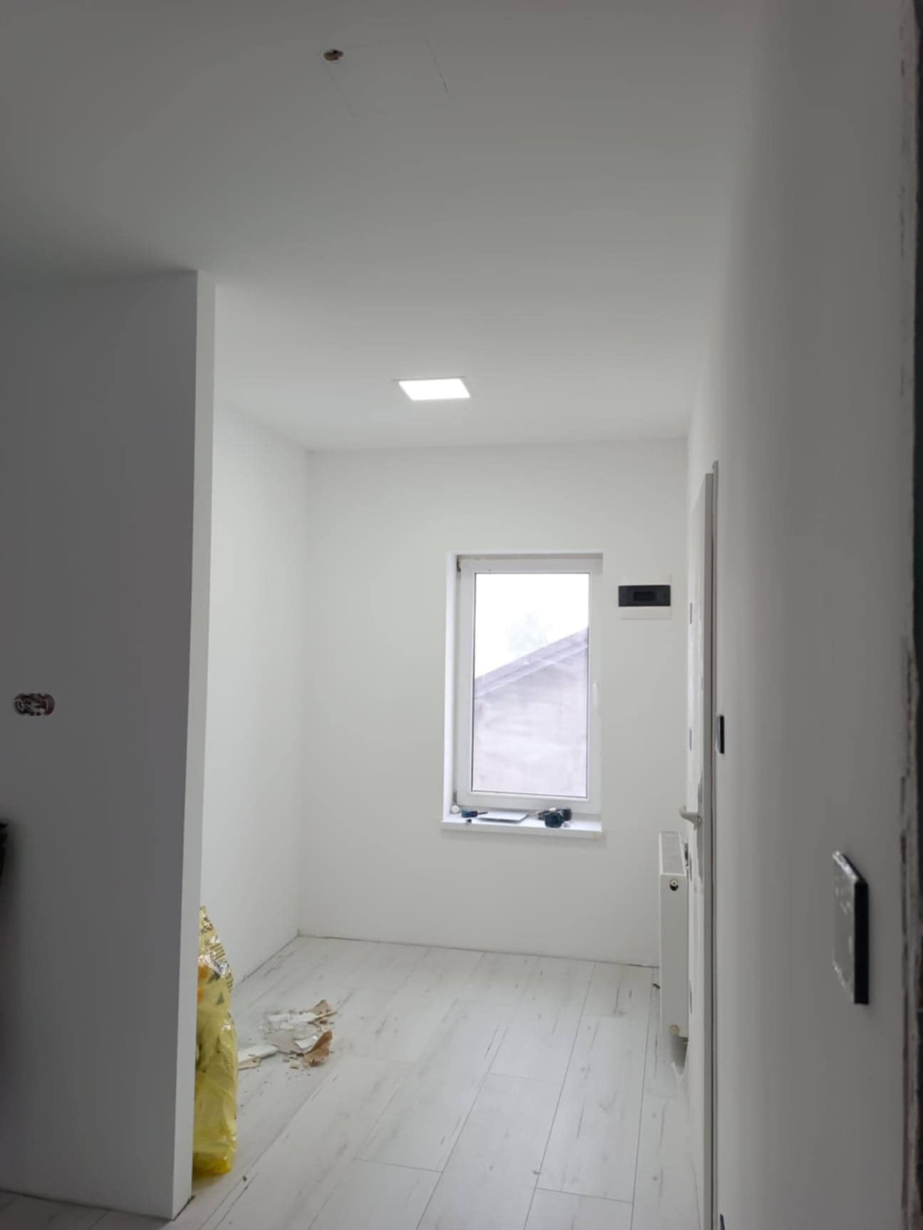 Wnętrze po remoncie: białe ściany, jasna podłoga, okno z widokiem na zewnątrz. Widoczne ślady po pracach wykończeniowych i grzejnik.