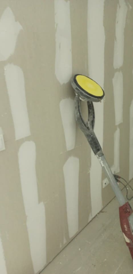 Szlifowanie ściany gipsowej szlifierką z żółtą tarczą. Widoczne paski szpachli. Praca w trakcie wygładzania powierzchni przed malowaniem.