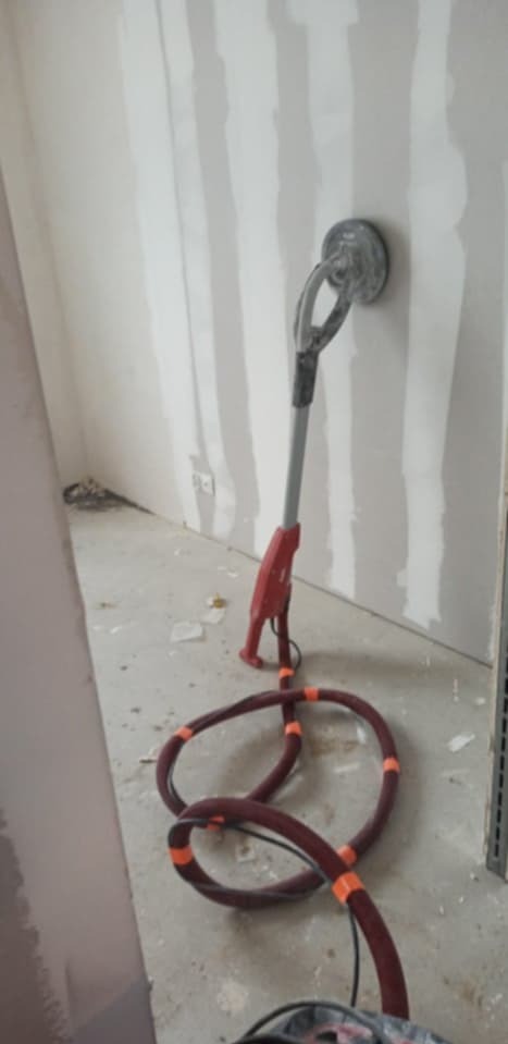 Szlifierka do gładzi gipsowych przy ścianie z widocznymi śladami szpachlowania, przygotowana do szlifowania. Widoczny przewód odkurzacza i fragment podłogi.