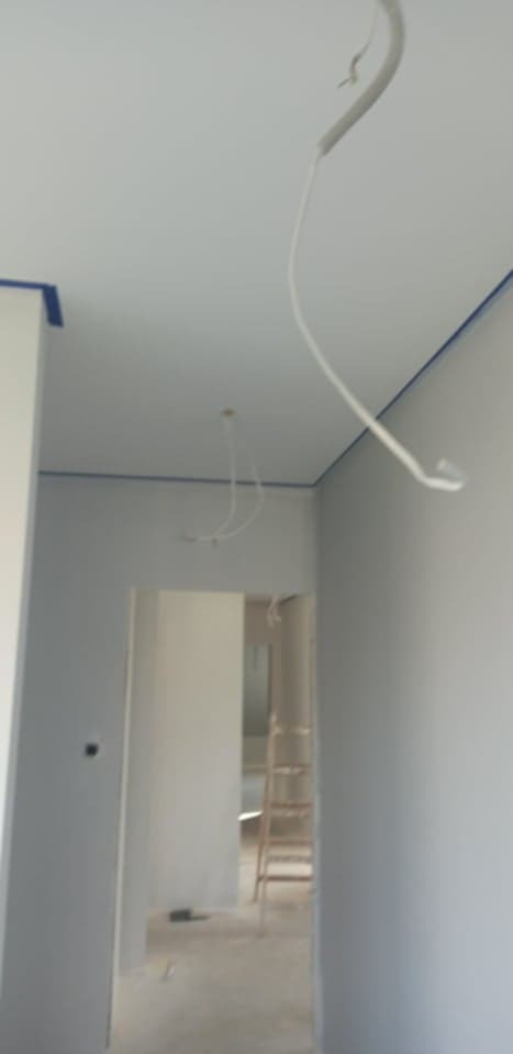 Wnętrze w trakcie malowania: świeżo pomalowane ściany na szaro, niebieska taśma malarska przy suficie, widoczne kable oświetleniowe i drabina w głębi pomieszczenia.