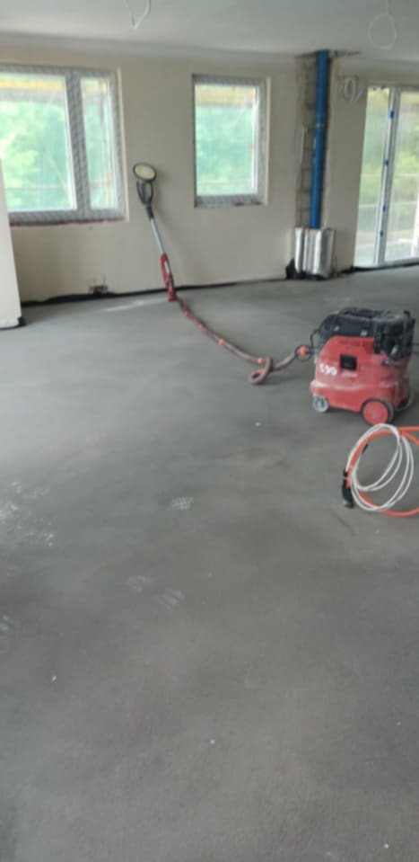 Wyrównywanie wylewki samopoziomującej za pomocą szlifierki i odkurzacza przemysłowego w pomieszczeniu w Kazimierzu. Widoczne okna i instalacje.