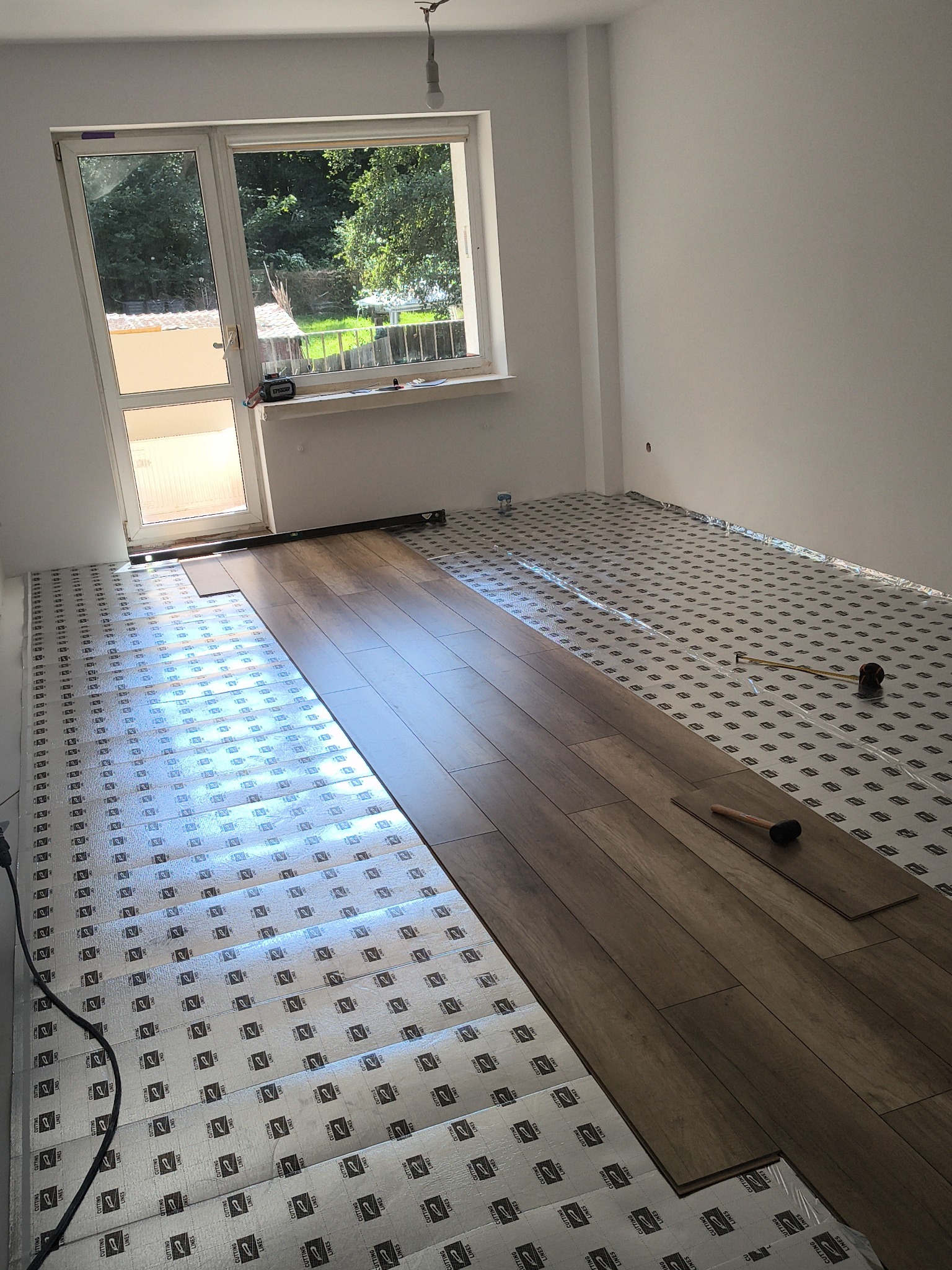 Układanie paneli podłogowych w jasnym pokoju z oknem. Widoczne narzędzia i podkład pod panele. Częściowo ułożona podłoga z paneli w odcieniach brązu.