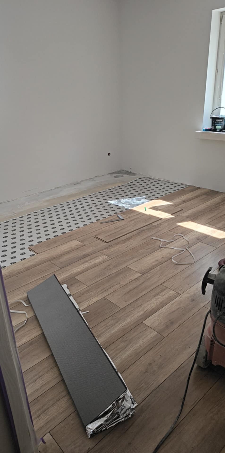 Układanie paneli podłogowych w jasnym pokoju. Widoczny podkład i częściowo ułożone panele. W tle ściana z oknem i narzędzia pracy.