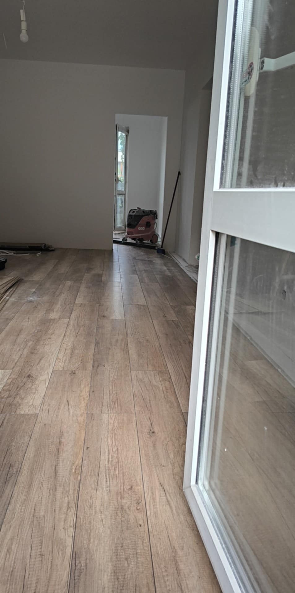 Ułożona podłoga z paneli w jasnym odcieniu drewna, widoczny odkurzacz przemysłowy i częściowo okno. Remont mieszkania w trakcie wykończenia.