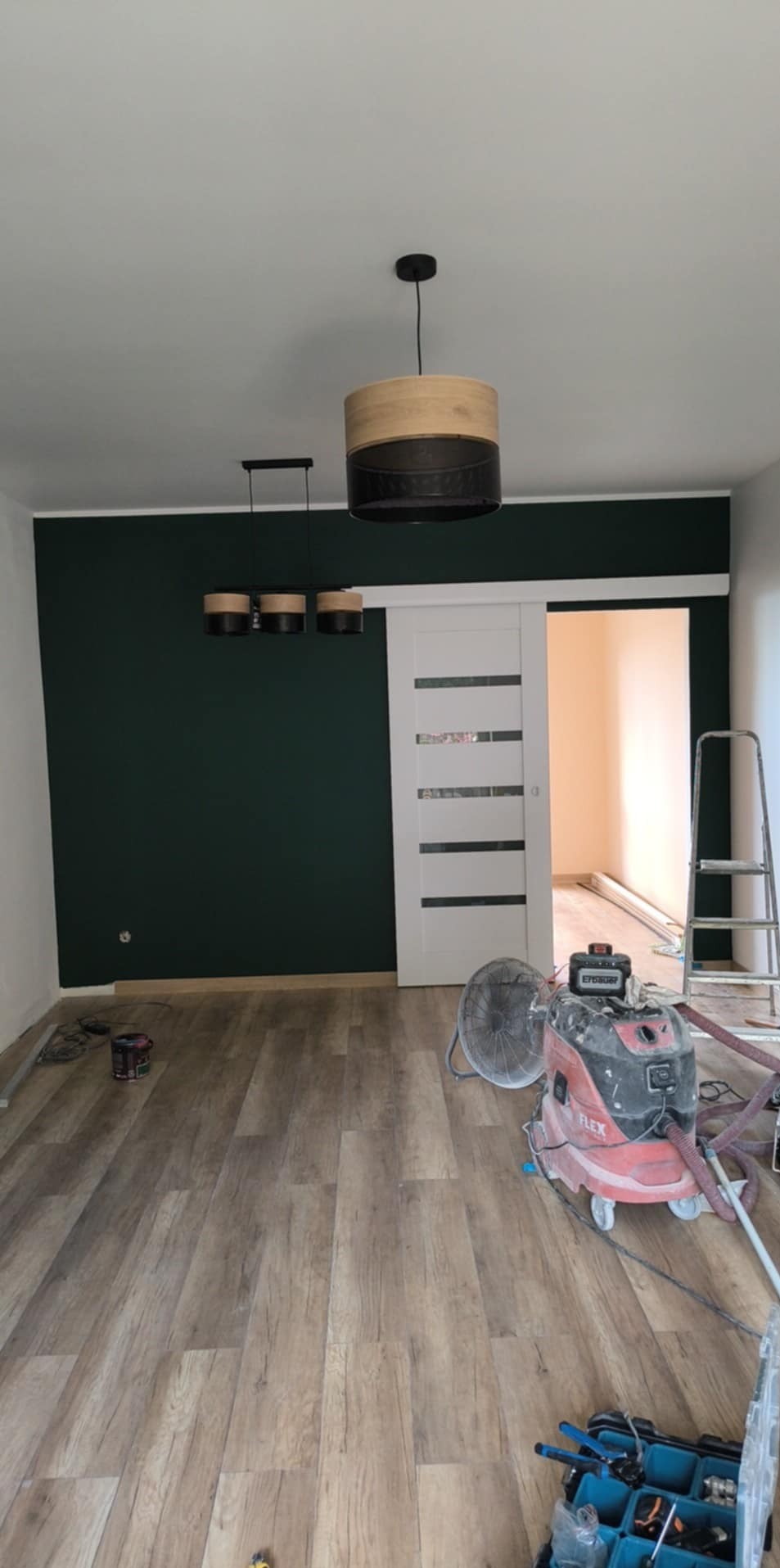 Pokój po malowaniu ściany na ciemnozielono i montażu przesuwnych drzwi. Widoczny odkurzacz przemysłowy, drabina i lampy. Podłoga z paneli imitujących drewno.