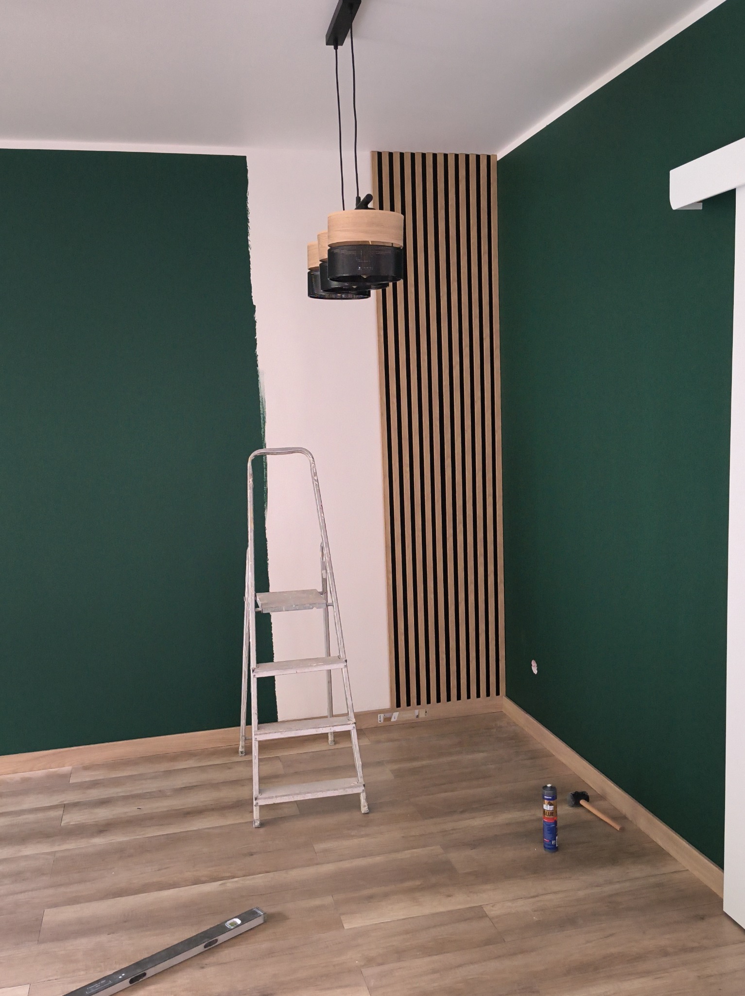 Wnętrze w trakcie malowania na butelkową zieleń z drewnianą lamelą na ścianie. Widoczna drabina, poziomica i narzędzia na podłodze. Częściowo pomalowana ściana.