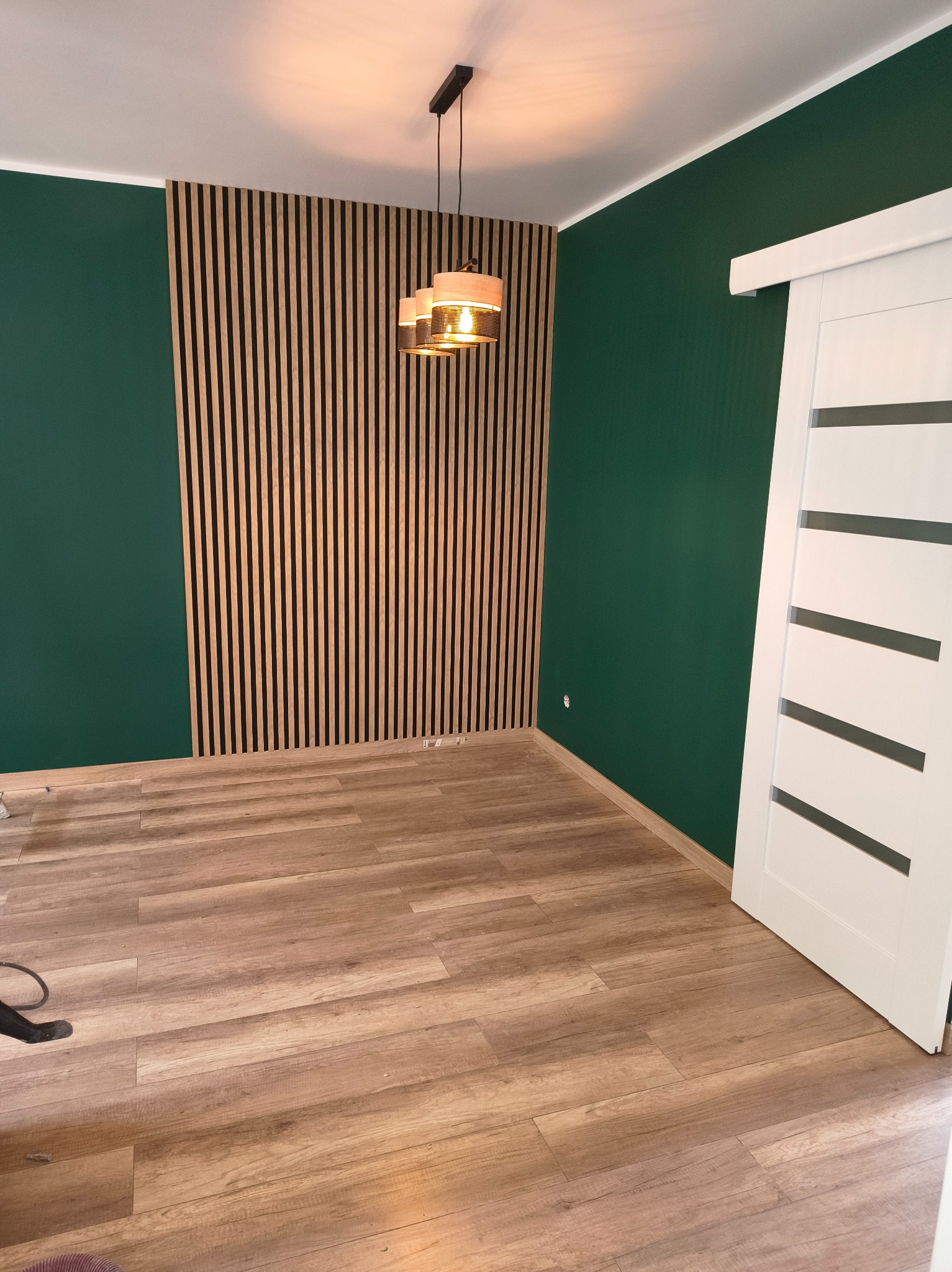 Nowoczesne wnętrze z zielonymi ścianami, drewnianą lamelą na ścianie i przesuwnymi białymi drzwiami. Widoczna drewniana podłoga i wisząca lampa.