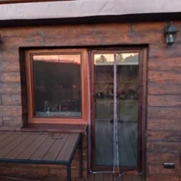 Drewniana elewacja domu z oknem i drzwiami balkonowymi, widoczne lampy zewnętrzne. W oknach odbija się zachód słońca, a w drzwiach wnętrze kuchni.