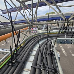 Krzysztof Kowalski - Rzędy czarnych kabli elektrycznych, starannie ułożonych na metalowej konstrukcji wsporczej. Kable zabezpieczone czarnymi opaskami. Widok z góry, na zewnątrz.