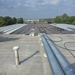Krzysztof Kowalski - Instalacja koryt kablowych na płaskim dachu budynku przemysłowego w słoneczny dzień. Widoczne elementy wentylacji i konstrukcja dachu z blachy trapezowej.