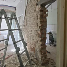 Demontaż ściany z cegły podczas remontu, widoczne fragmenty instalacji, drabina i gruz. Prace w toku, surowy stan wnętrza.