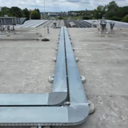 Szara, metalowa trasa kablowa na betonowym dachu z widocznymi panelami słonecznymi w tle, zamontowana na betonowych wspornikach. Widok z góry na instalację.