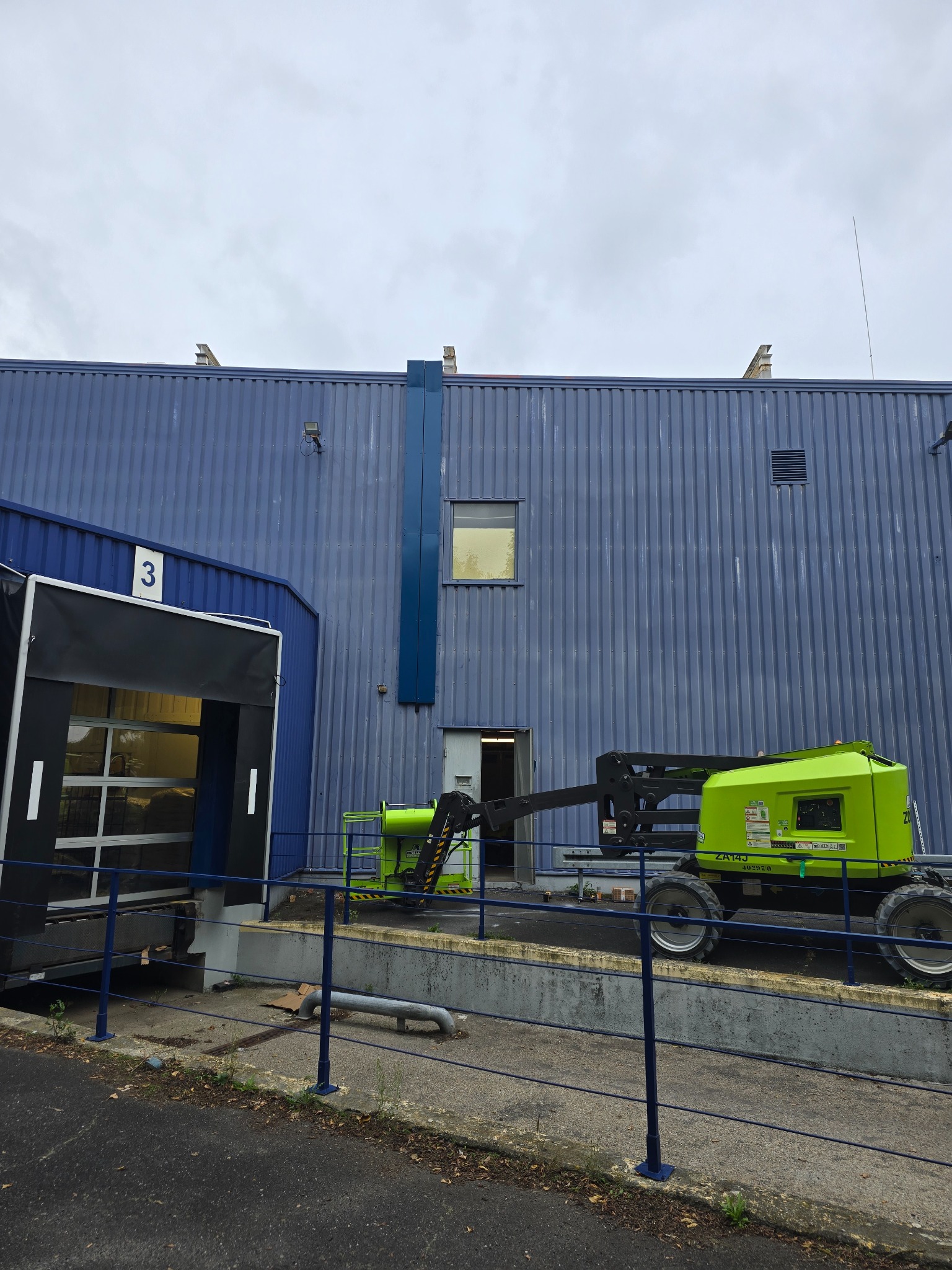 Zielony podnośnik koszowy na tle niebieskiej hali z numerem 3 przy rampie załadunkowej. Widoczny fragment ogrodzenia.