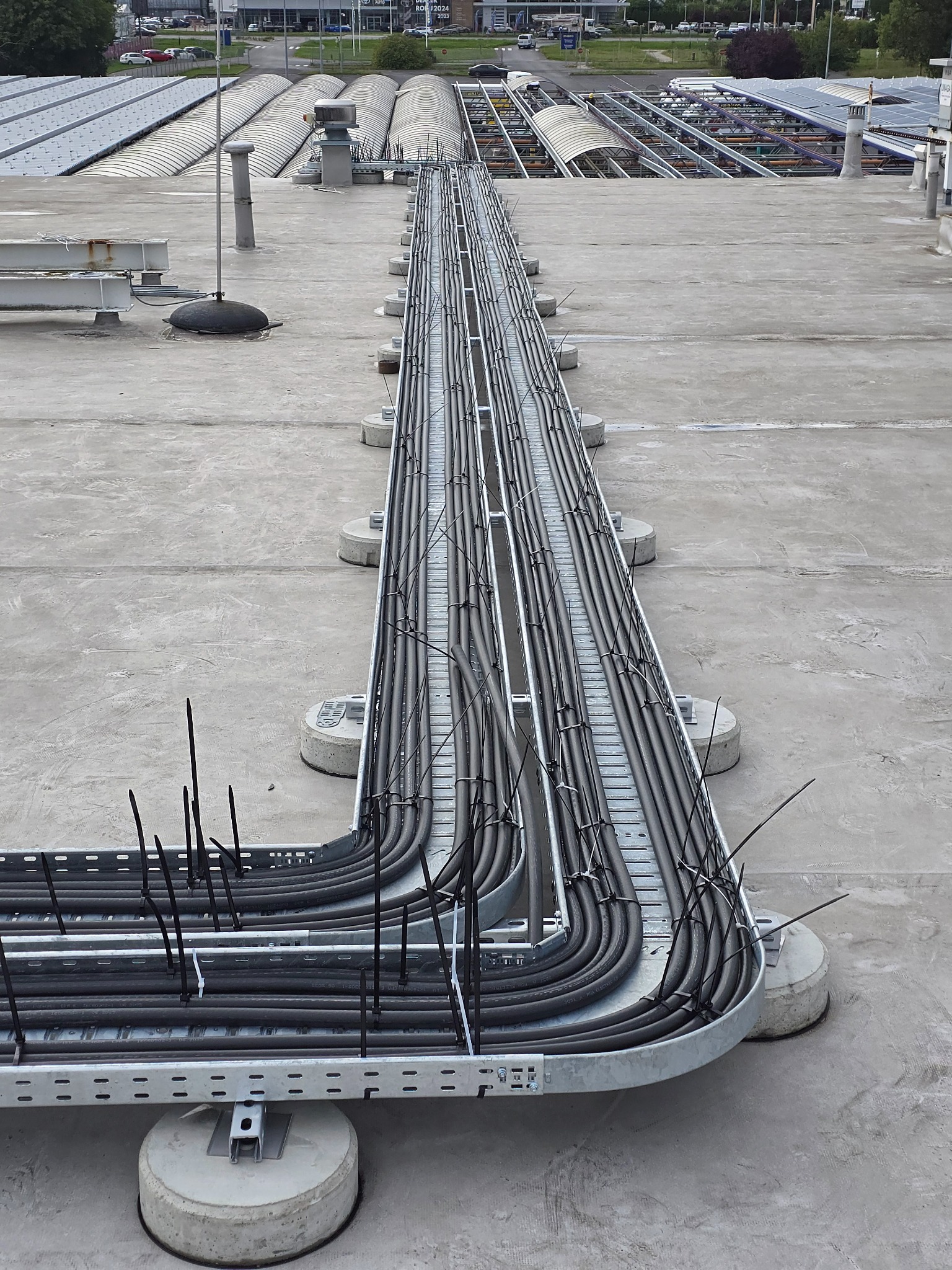 Instalacja kablowa na dachu budynku komercyjnego, kable ułożone w korytkach, zabezpieczone opaskami. Widok z góry, betonowe podłoże, elementy wentylacji w tle.