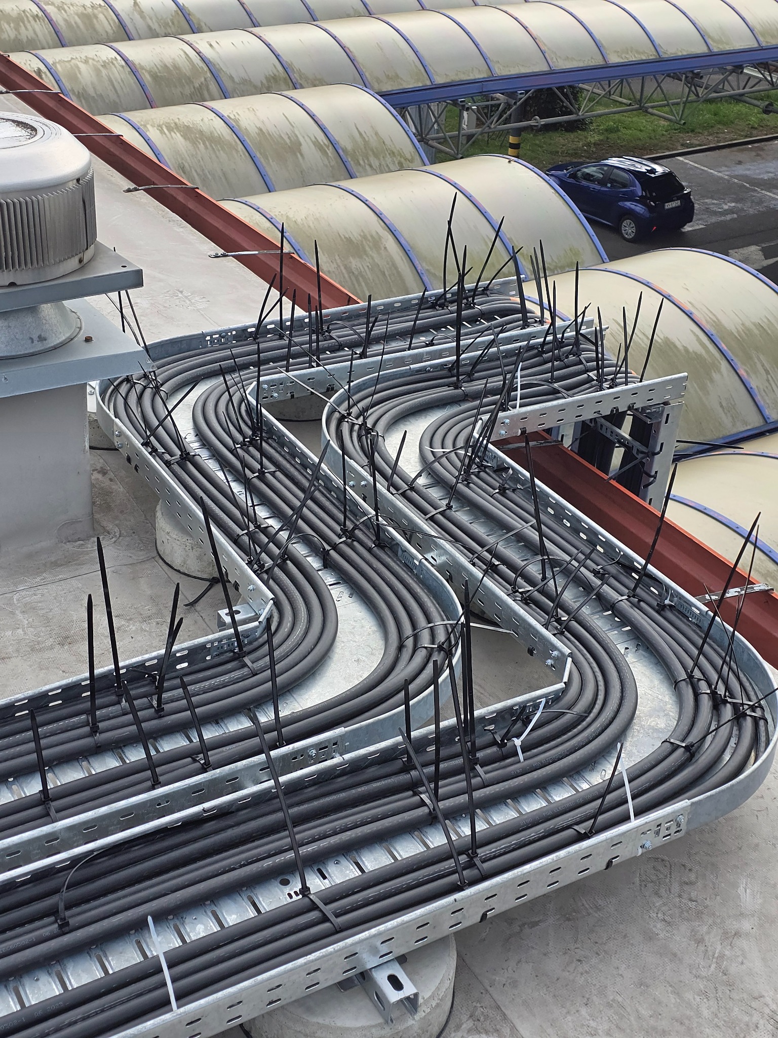 Zamocowane kable w metalowej korycie na dachu budynku, zabezpieczone czarnymi opaskami zaciskowymi. W tle widoczny fragment parkingu i zadaszenia.