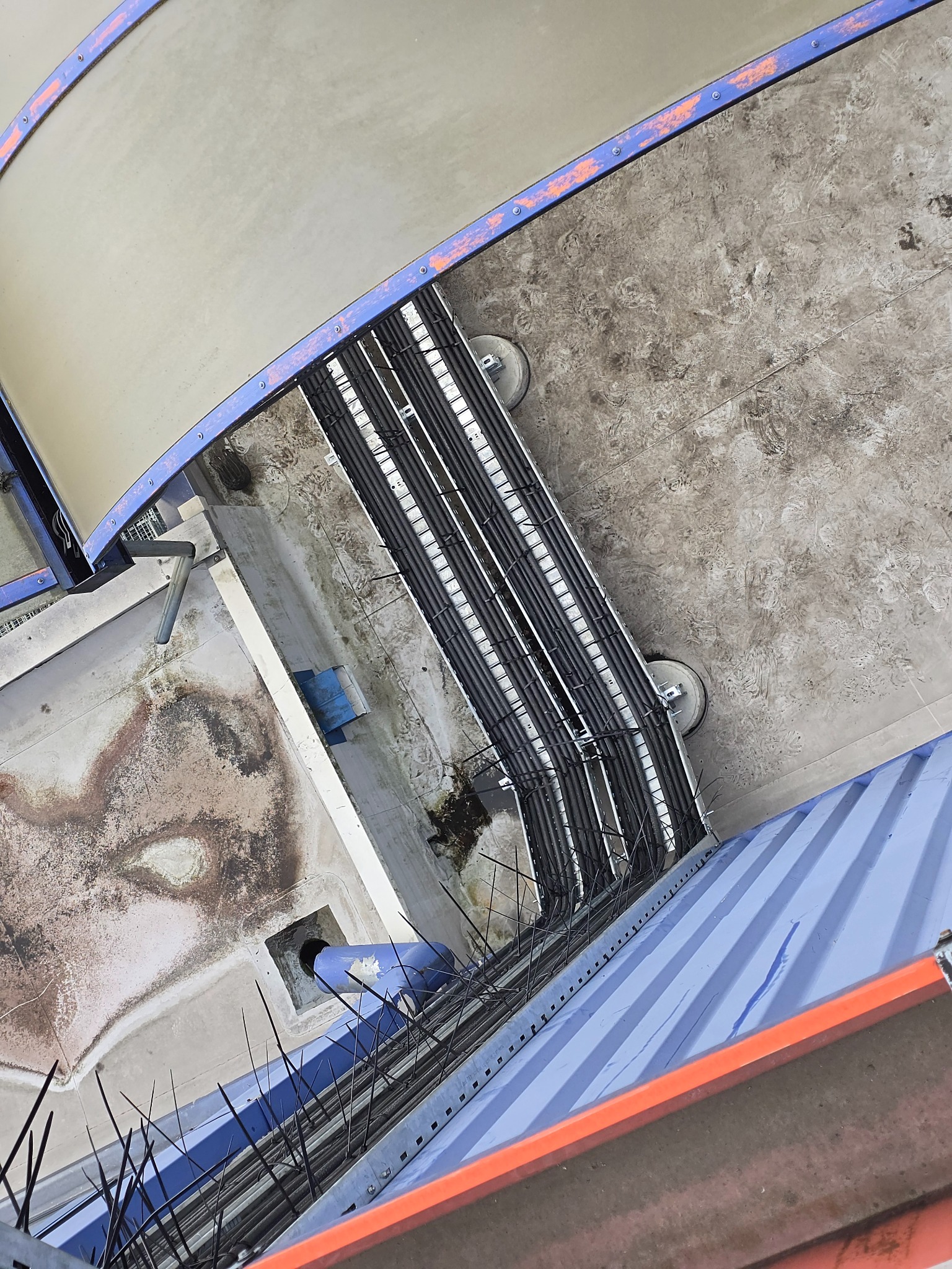 Widok z góry na instalacje rur w korycie kablowym, z elementami konstrukcyjnymi budynku. Rury ułożone równolegle, widoczne mocowania i zabezpieczenia.