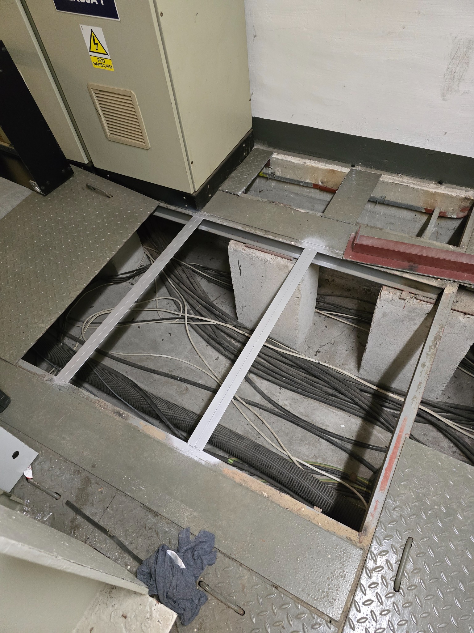 Otwór w podłodze technicznej z widocznymi kablami i przewodami. Metalowa podłoga z kratownicą i wystający fragment szafy elektrycznej. Ułożone kable w przestrzeni pod podłogą.
