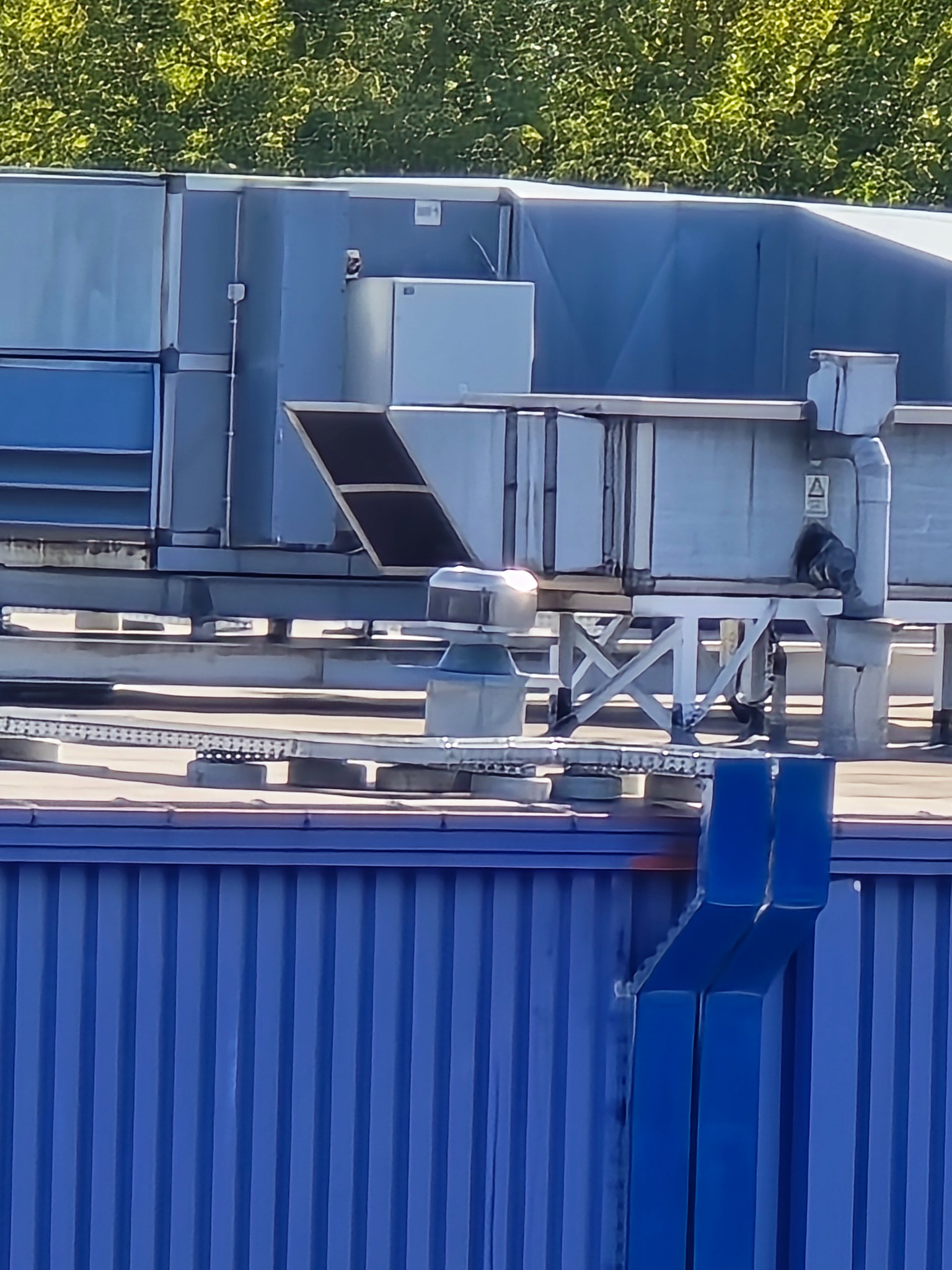 Kompleksowa instalacja wentylacyjna na dachu budynku z niebieską elewacją. Widoczne kanały, filtry i elementy mocujące. Ujęcie z dołu, w tle zieleń drzew.