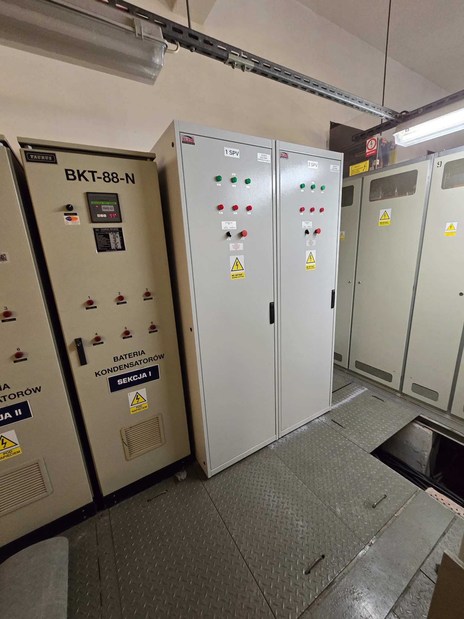 Rozdzielnia elektryczna z szafami sterowniczymi i baterią kondensatorów, widoczne kontrolki, oznaczenia ostrzegawcze i metalowa podłoga.