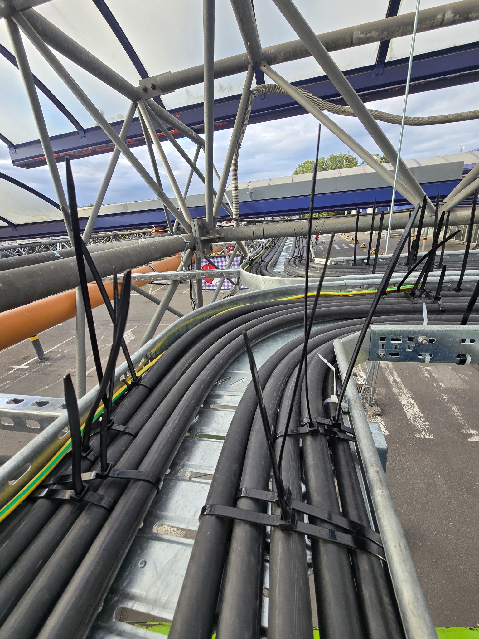 Rzędy czarnych kabli elektrycznych, starannie ułożonych na metalowej konstrukcji wsporczej. Kable zabezpieczone czarnymi opaskami. Widok z góry, na zewnątrz.