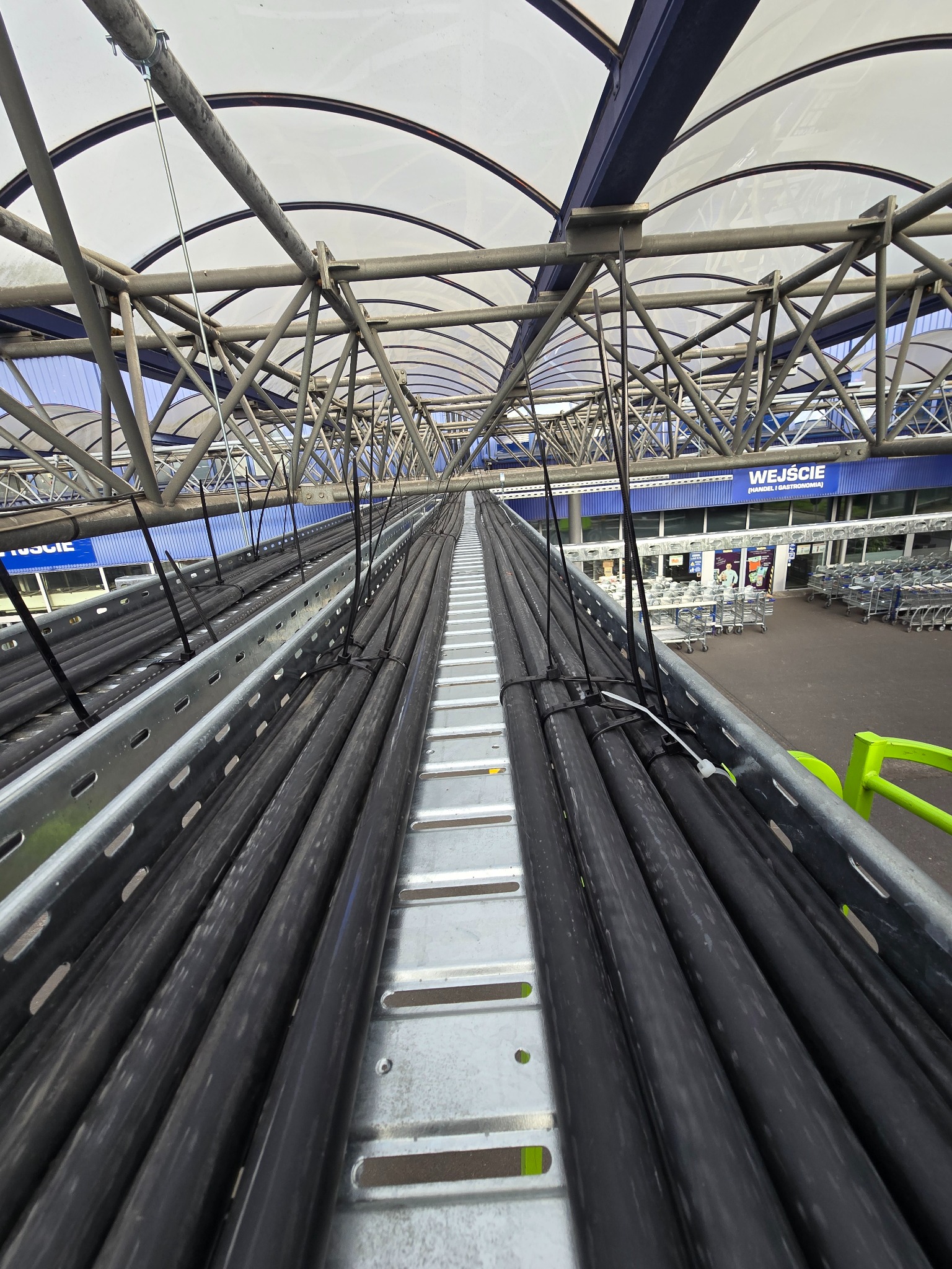 Rząd czarnych rur przymocowanych do metalowej konstrukcji pod zadaszeniem, widok z góry na zewnątrz budynku.