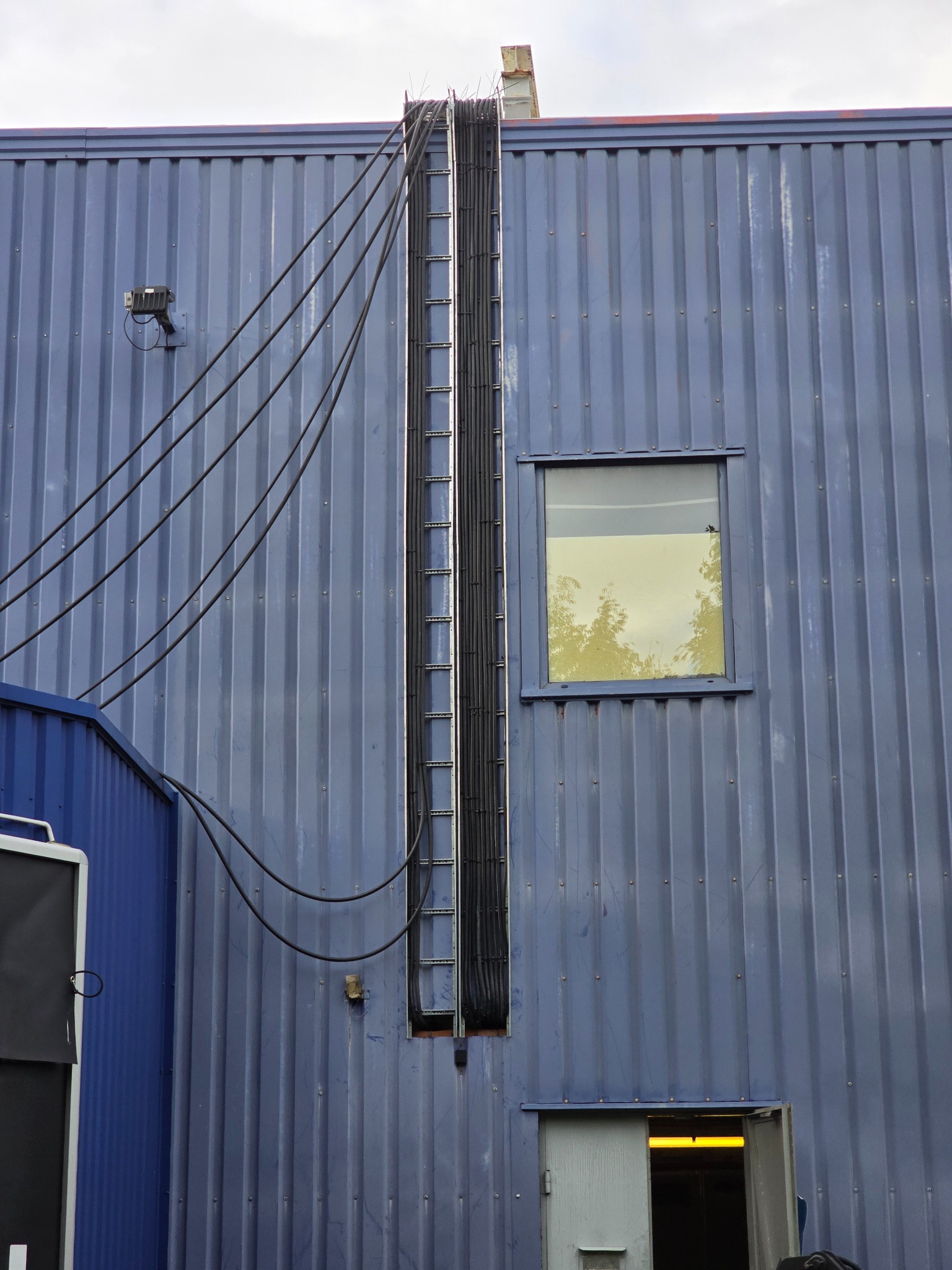 Zewnętrzna instalacja elektryczna na błękitnej, metalowej ścianie budynku. Widoczne kable, drabinka kablowa, okno i częściowo otwarte drzwi.