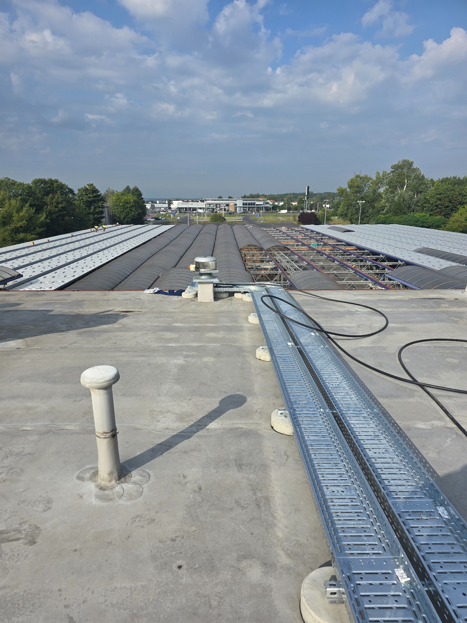 Instalacja koryt kablowych na płaskim dachu budynku przemysłowego w słoneczny dzień. Widoczne elementy wentylacji i konstrukcja dachu z blachy trapezowej.