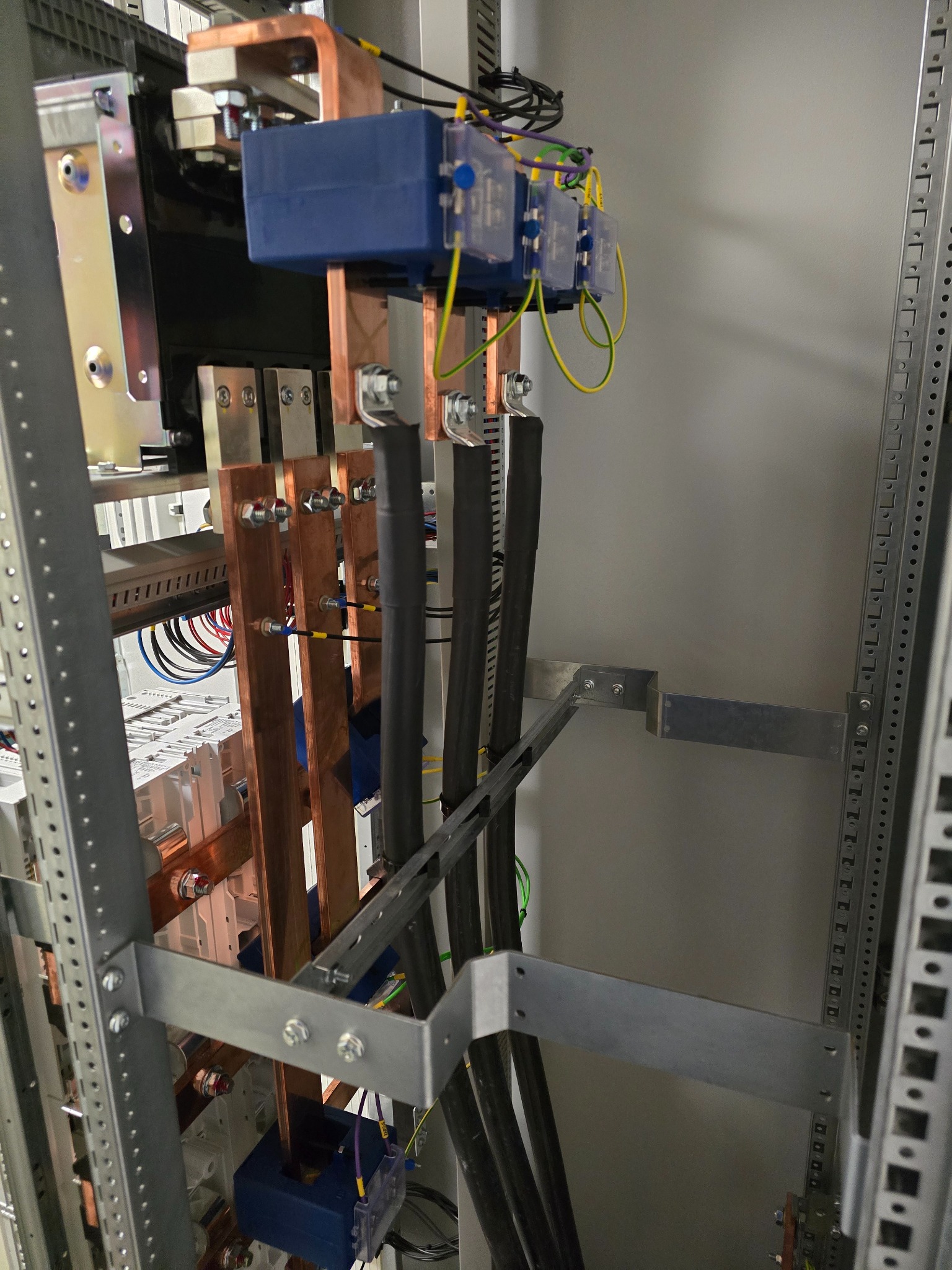 Rozdzielnica elektryczna z miedzianymi szynami, bezpiecznikami i czarnymi kablami, zamontowana wewnątrz metalowej szafy, widok z bliska na instalację.