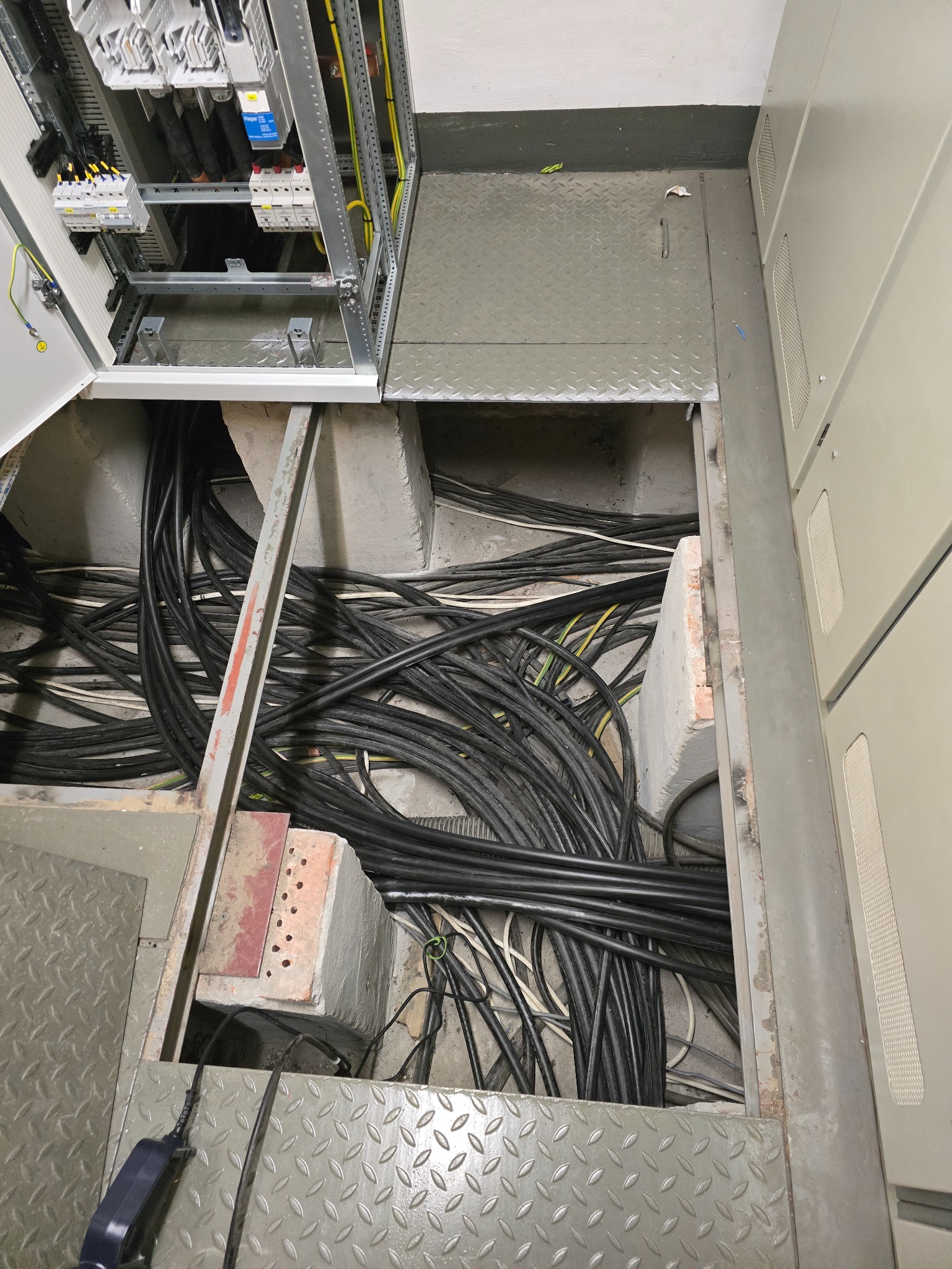 Rozłożone kable w podłodze technicznej, widoczne elementy szafy elektrycznej w Bydgoszczy. Prace instalacyjne w toku, widok z góry na otwartą przestrzeń pod podłogą.