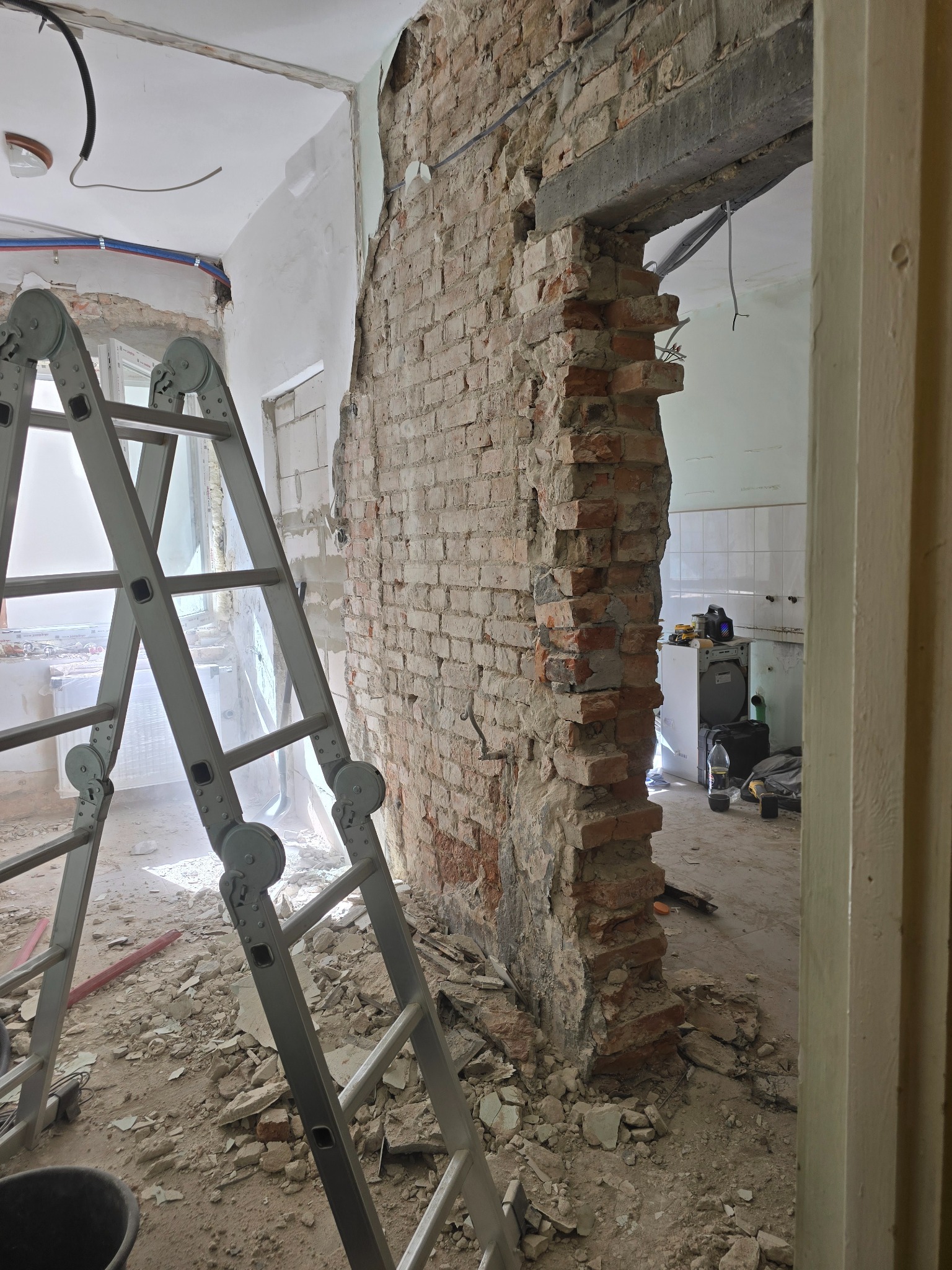Demontaż ściany z cegły podczas remontu, widoczne fragmenty instalacji, drabina i gruz. Prace w toku, surowy stan wnętrza.