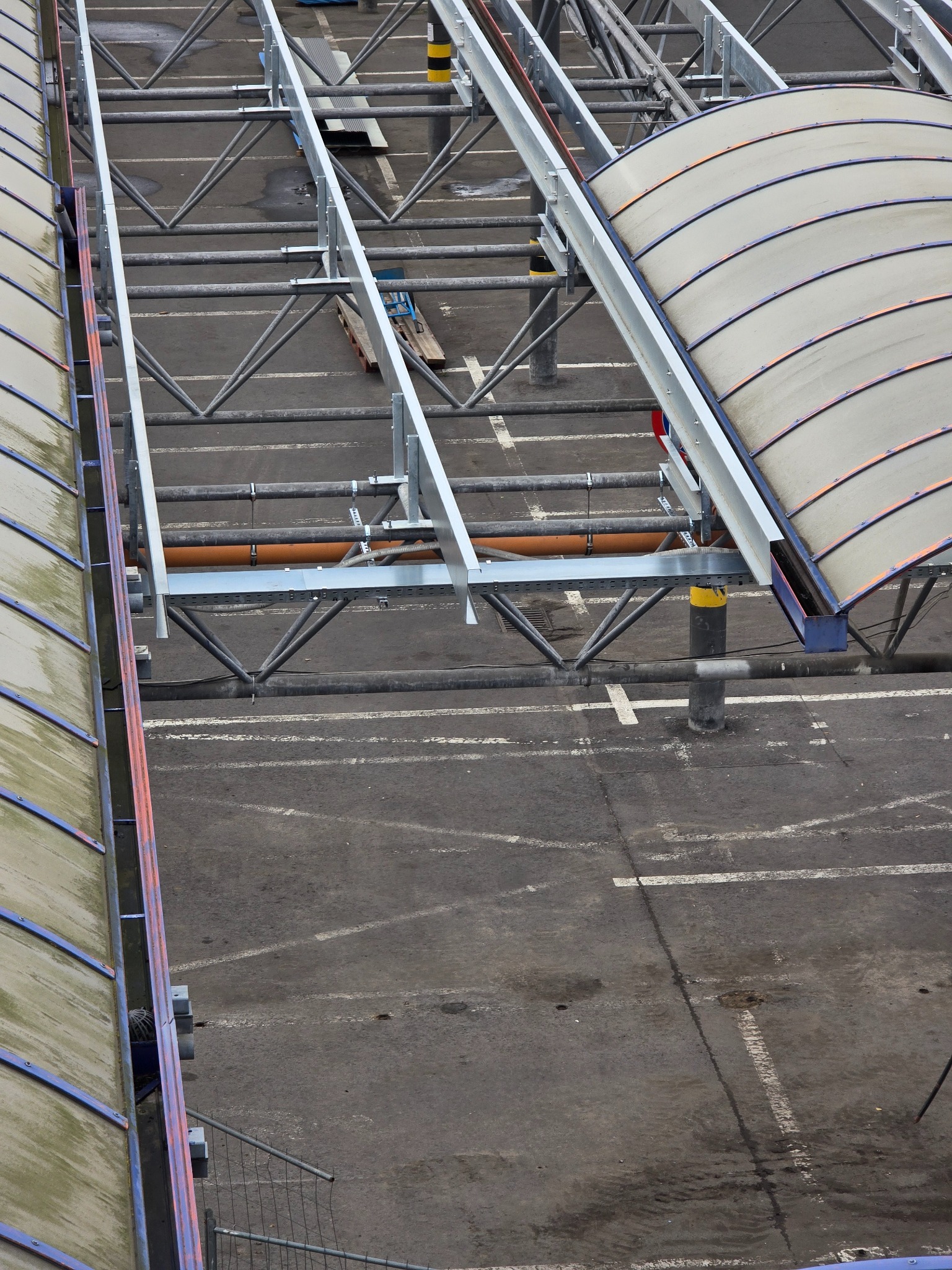 Z góry widok na stalową konstrukcję z zadaszeniem na parkingu, z widocznymi rurami i elementami montażowymi. Szare tło asfaltu z białymi liniami parkingowymi.