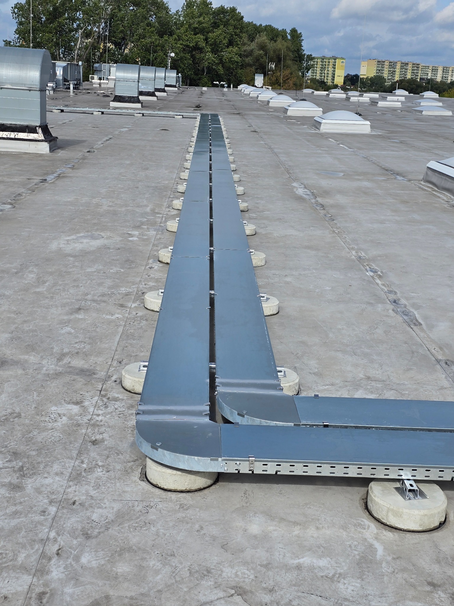 Szary, metalowy system kanałów wentylacyjnych na płaskim dachu budynku z widocznymi świetlikami i budynkami w tle, wykonany z dbałością o detale.