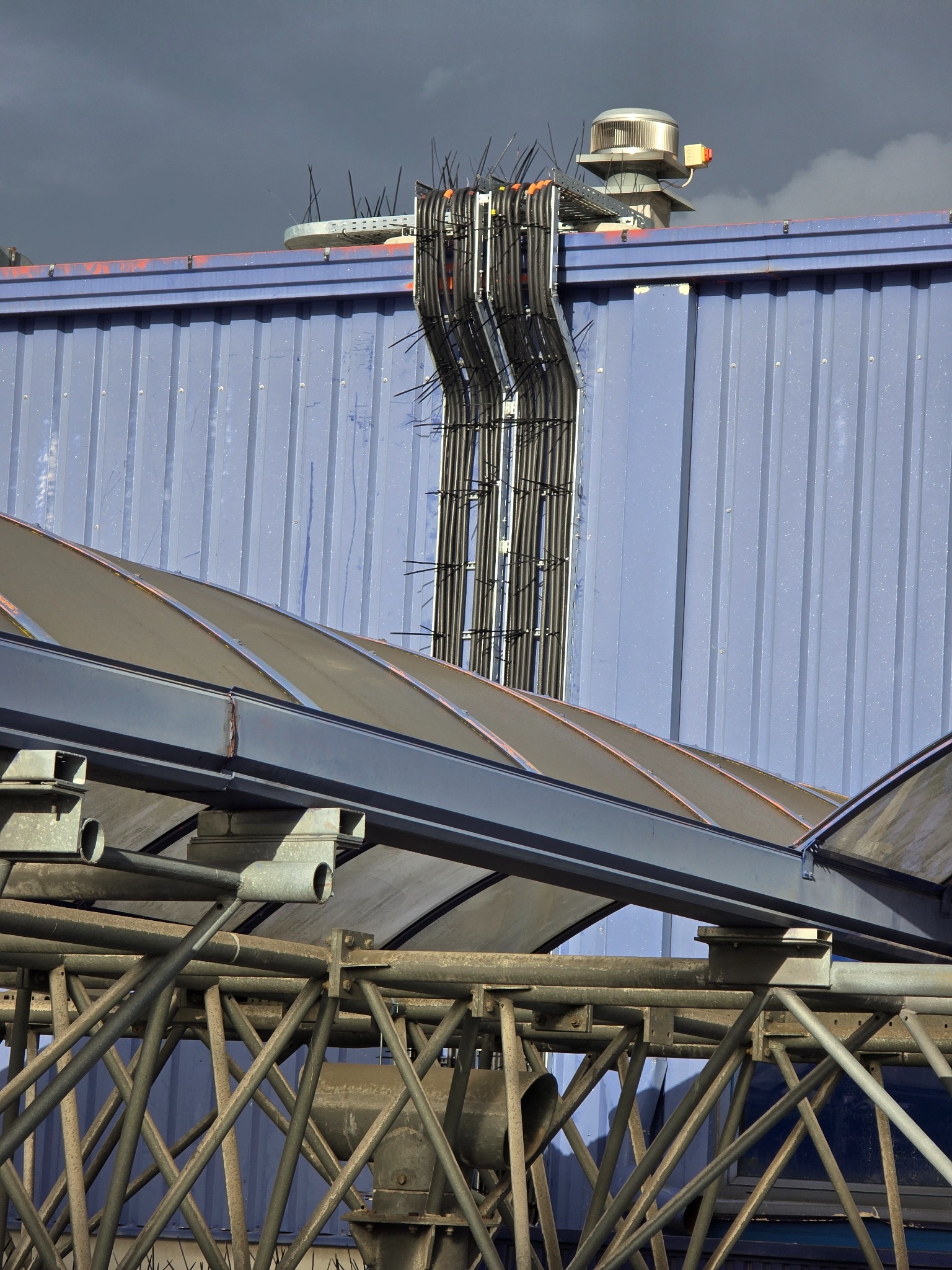 Zewnętrzna instalacja elektryczna na błękitnej, blaszanej ścianie budynku. Widoczne kable prowadzone po elewacji, metalowa konstrukcja dachu i wentylator na dachu.