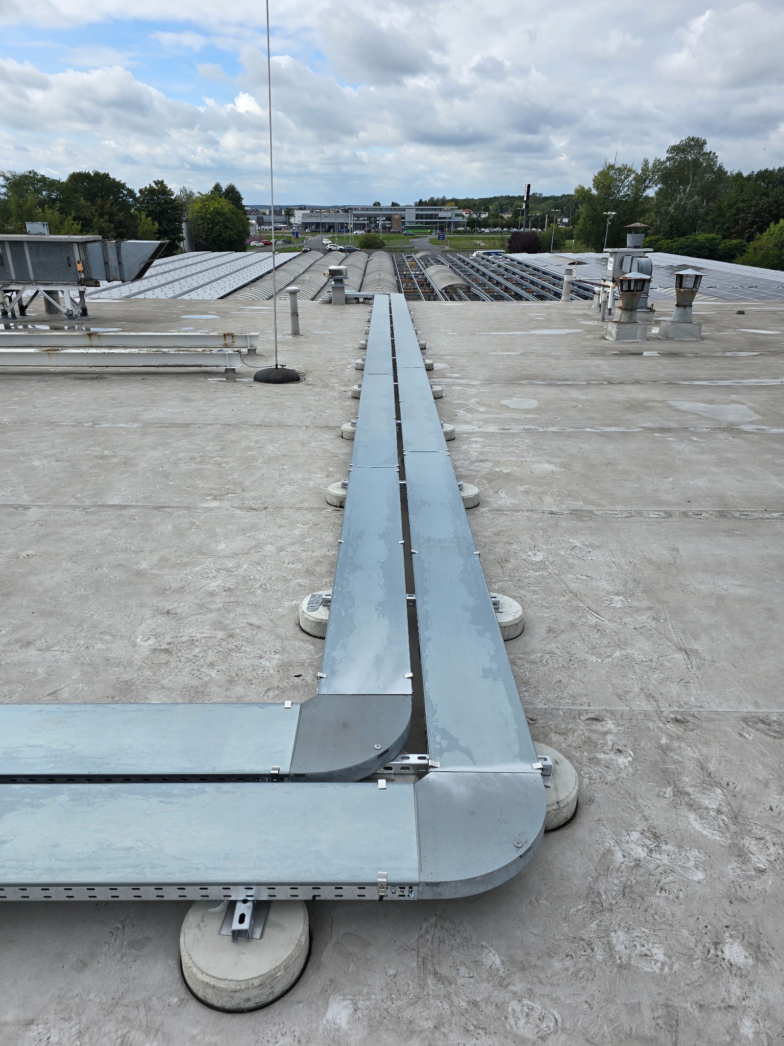 Szara, metalowa trasa kablowa na betonowym dachu z widocznymi panelami słonecznymi w tle, zamontowana na betonowych wspornikach. Widok z góry na instalację.