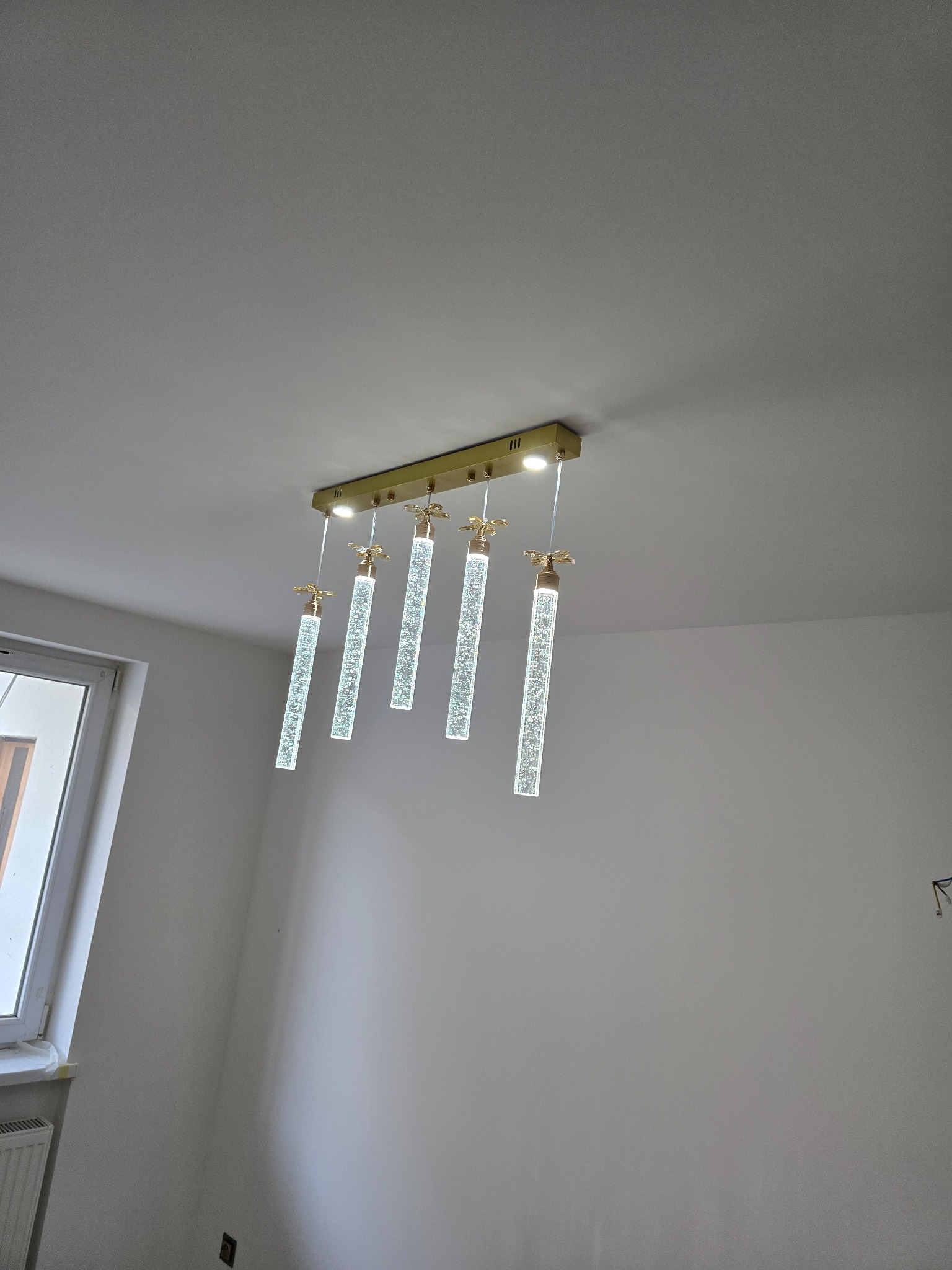 Nowoczesna lampa sufitowa z pięcioma szklanymi, podłużnymi kloszami z efektem bąbelków, zawieszona w jasnym wnętrzu, widok z dołu.