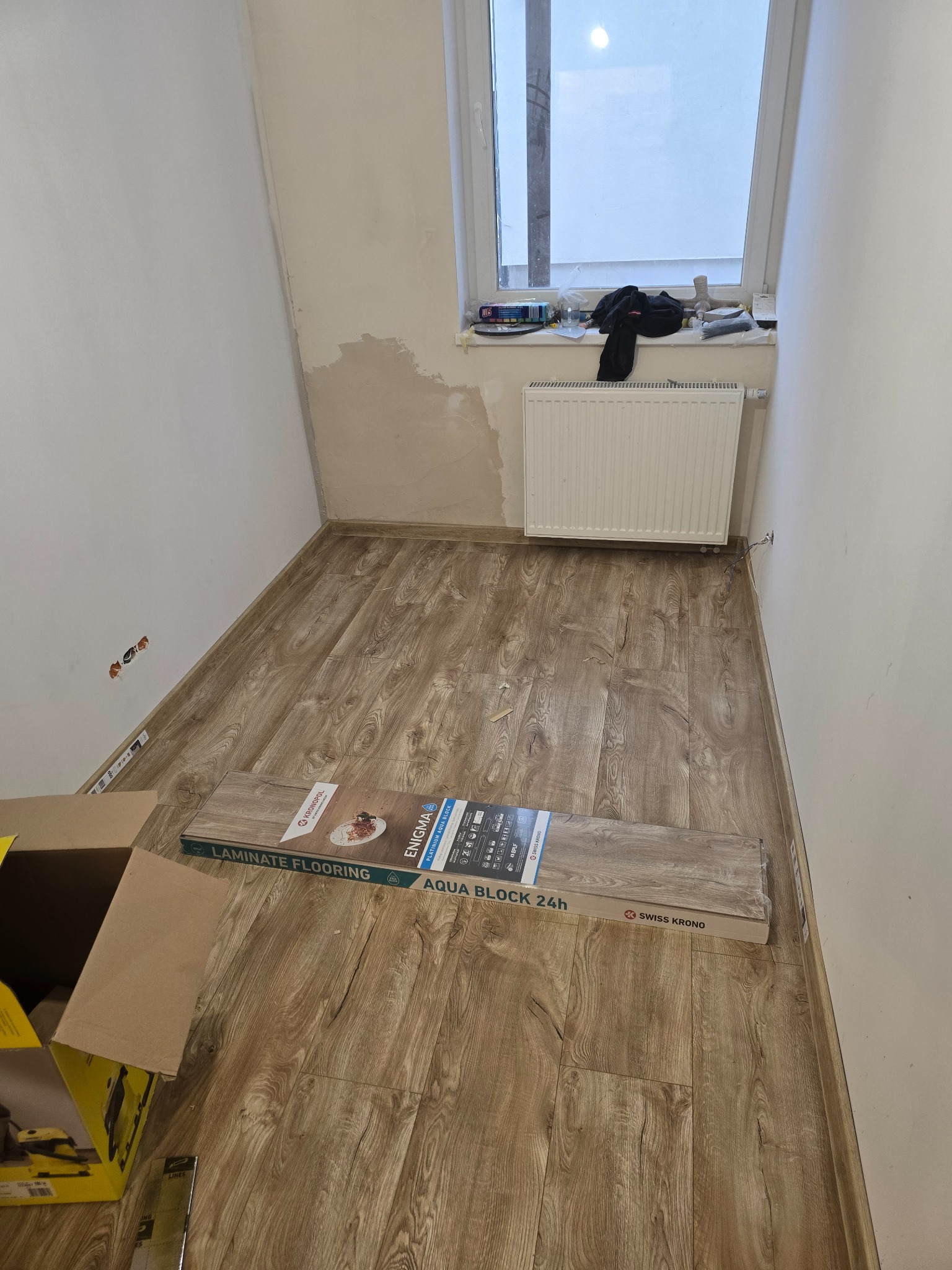 Ułożone panele podłogowe w małym pokoju, widoczny grzejnik i okno. Na podłodze leżą paczki paneli Swiss Krono Enigma Aqua Block 24h.