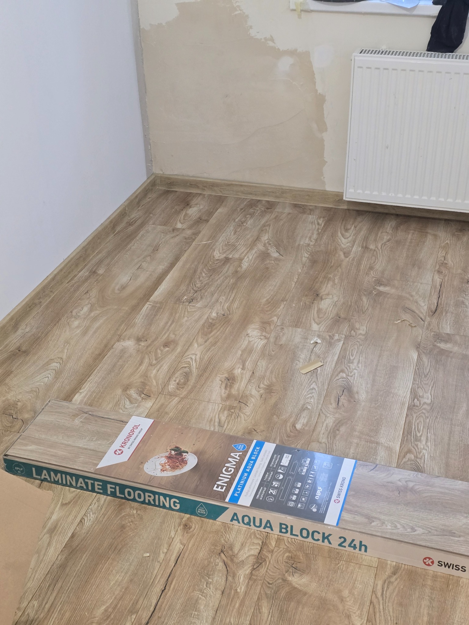 Ułożone panele podłogowe w pokoju, widoczny fragment ściany z grzejnikiem i paczka paneli Kronopol Enigma Aqua Block 24h. Jasne drewno, świeżo po montażu.