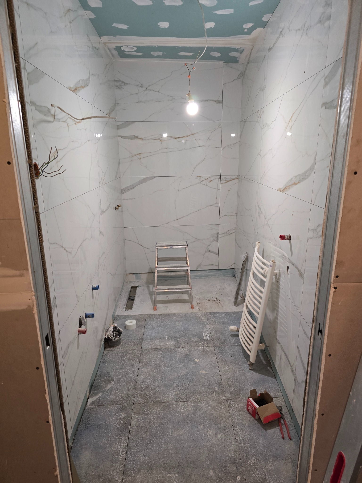 Remont łazienki w trakcie: białe płytki imitujące marmur na ścianach, szare płytki na podłodze, drabina, grzejnik, instalacja elektryczna i sufit z płyt kartonowo-gipsowych.